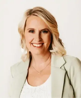 Blonde woman with a friendly smile, wearing a blazer and necklace, against a white background.