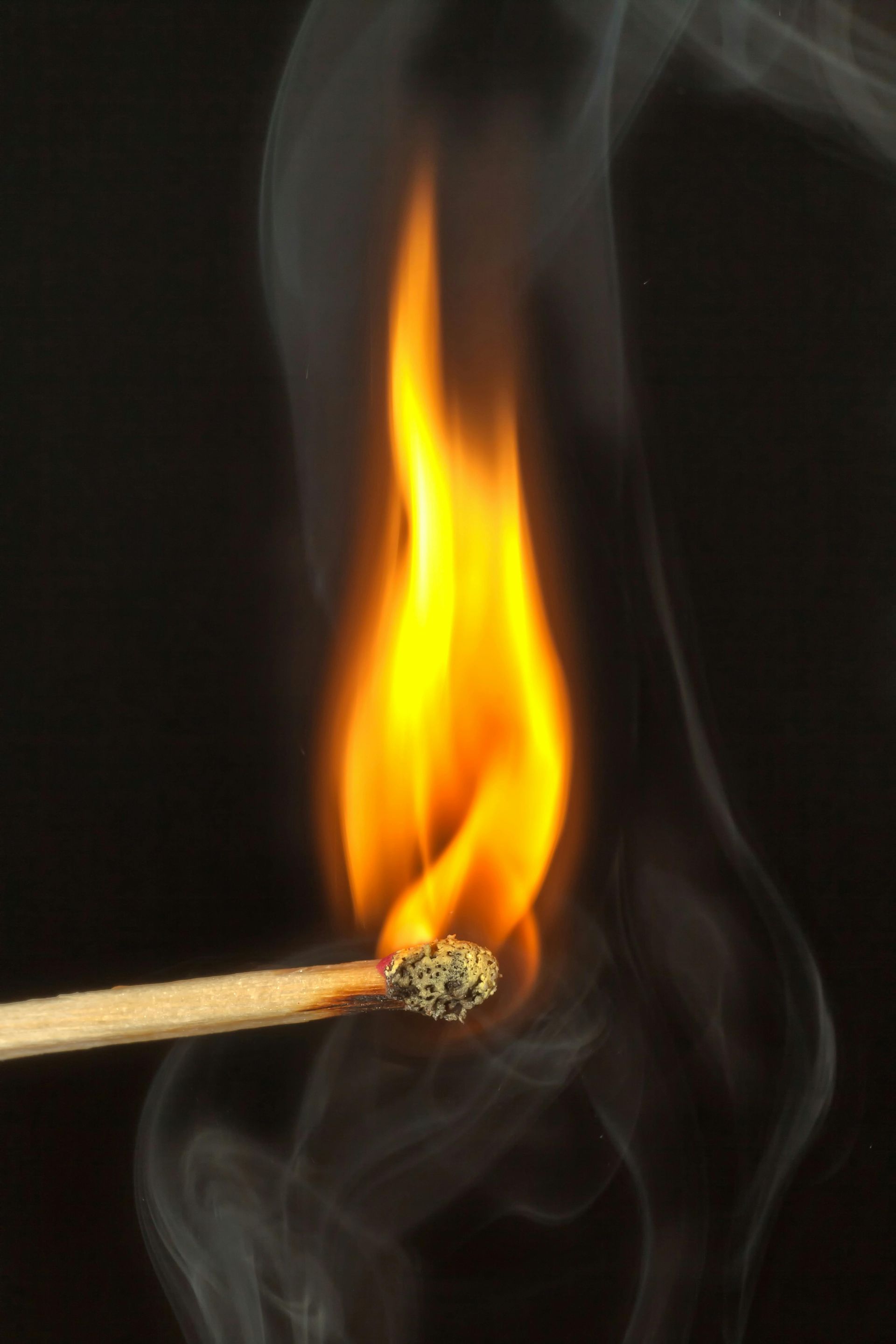 Burning matchstick with a bright yellow and orange flame, producing wisps of gray smoke against a black background.