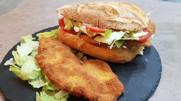 Panino con cotoletta impanata, lattuga e pomodoro su un piatto nero, accompagnato da una cotoletta e un'insalata a parte.