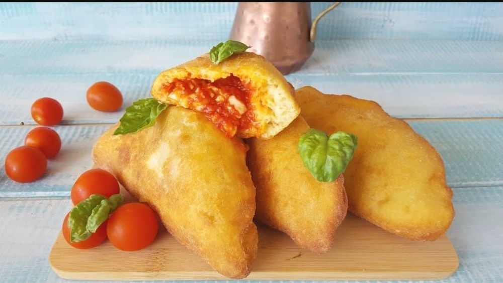 Tasche di pizza fritte con ripieno di pomodoro, guarnite con basilico e pomodori su un tagliere di legno.