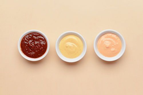 Tre piccole ciotole bianche con salsa rossa, gialla e arancione su una superficie beige.