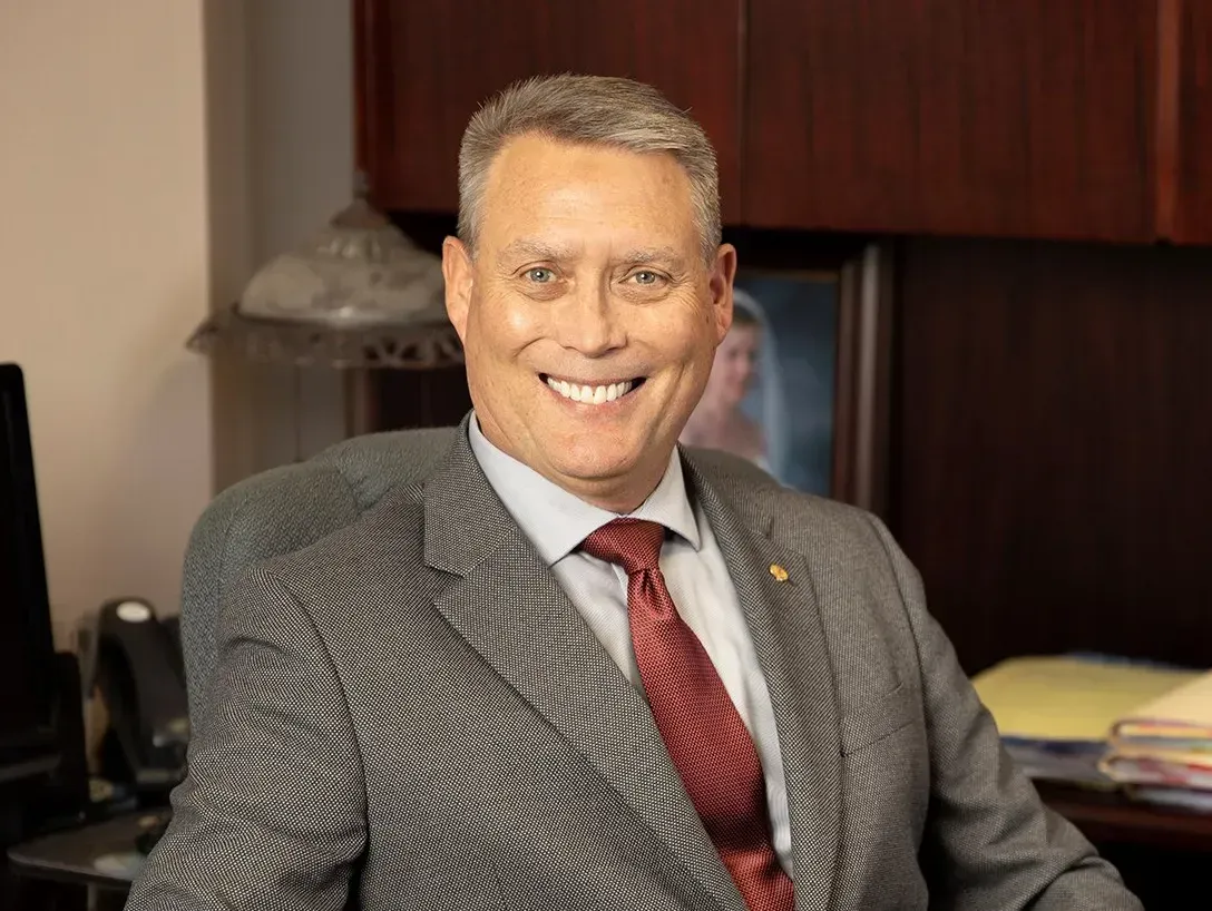 A smiling person in a grey blazer and red patterned tie sitting in an office setting.