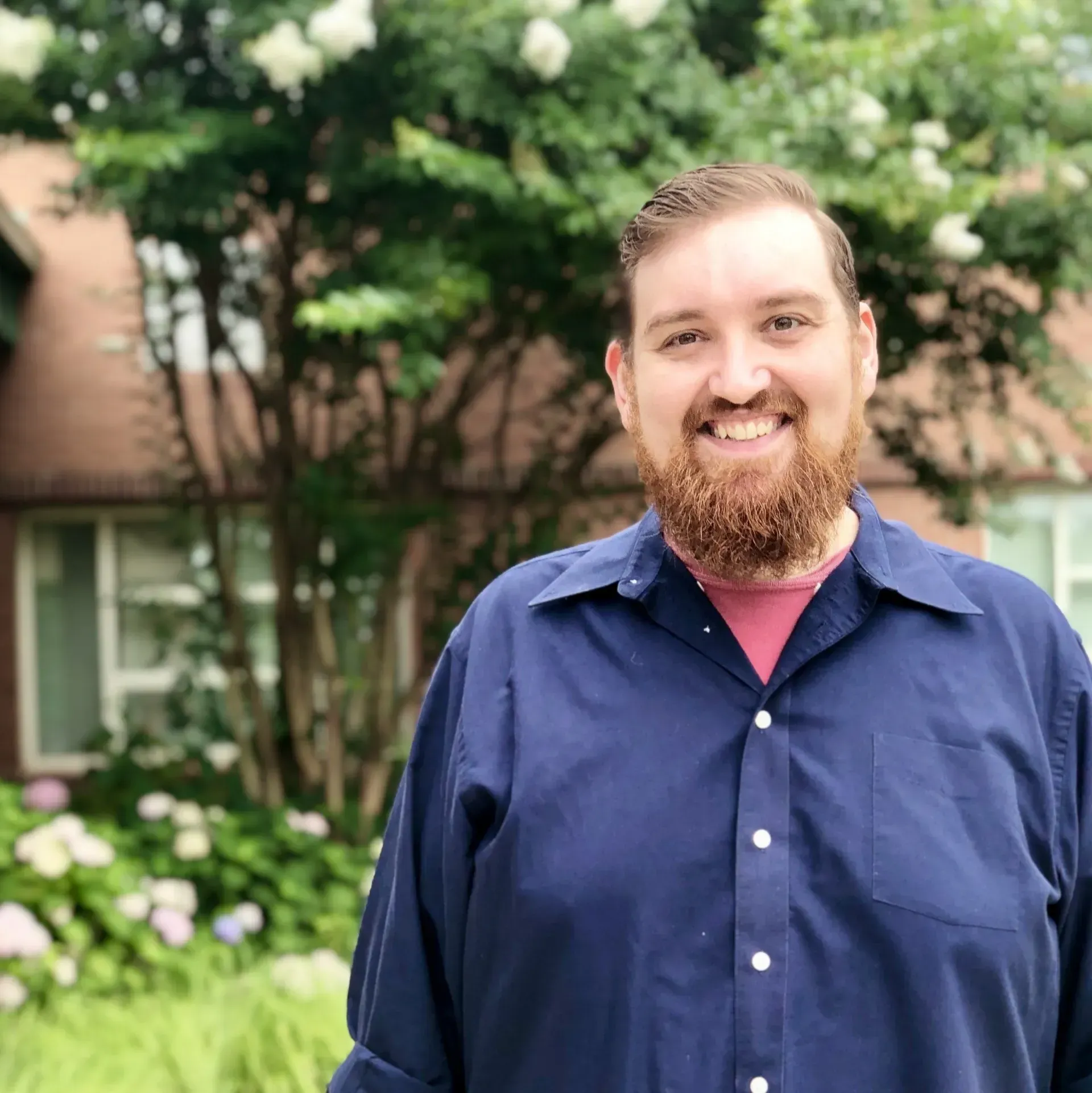 A smiling person with a beard, wearing a blue button-down shirt over a pink undershirt, posing outdoors near a tree.