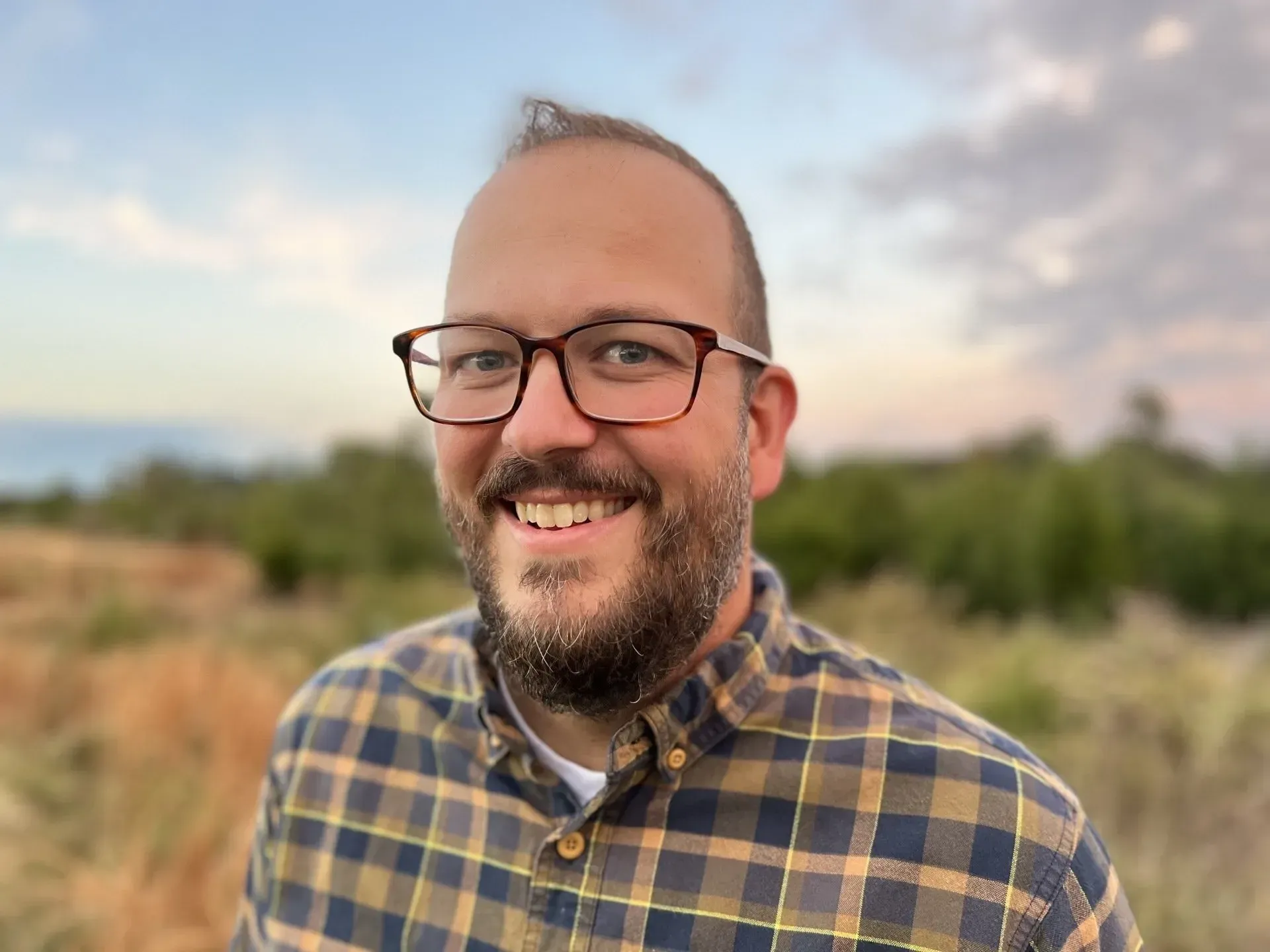 A person wearing glasses and a plaid shirt smiles outdoors in a field with a soft, sunset-lit sky in the background.