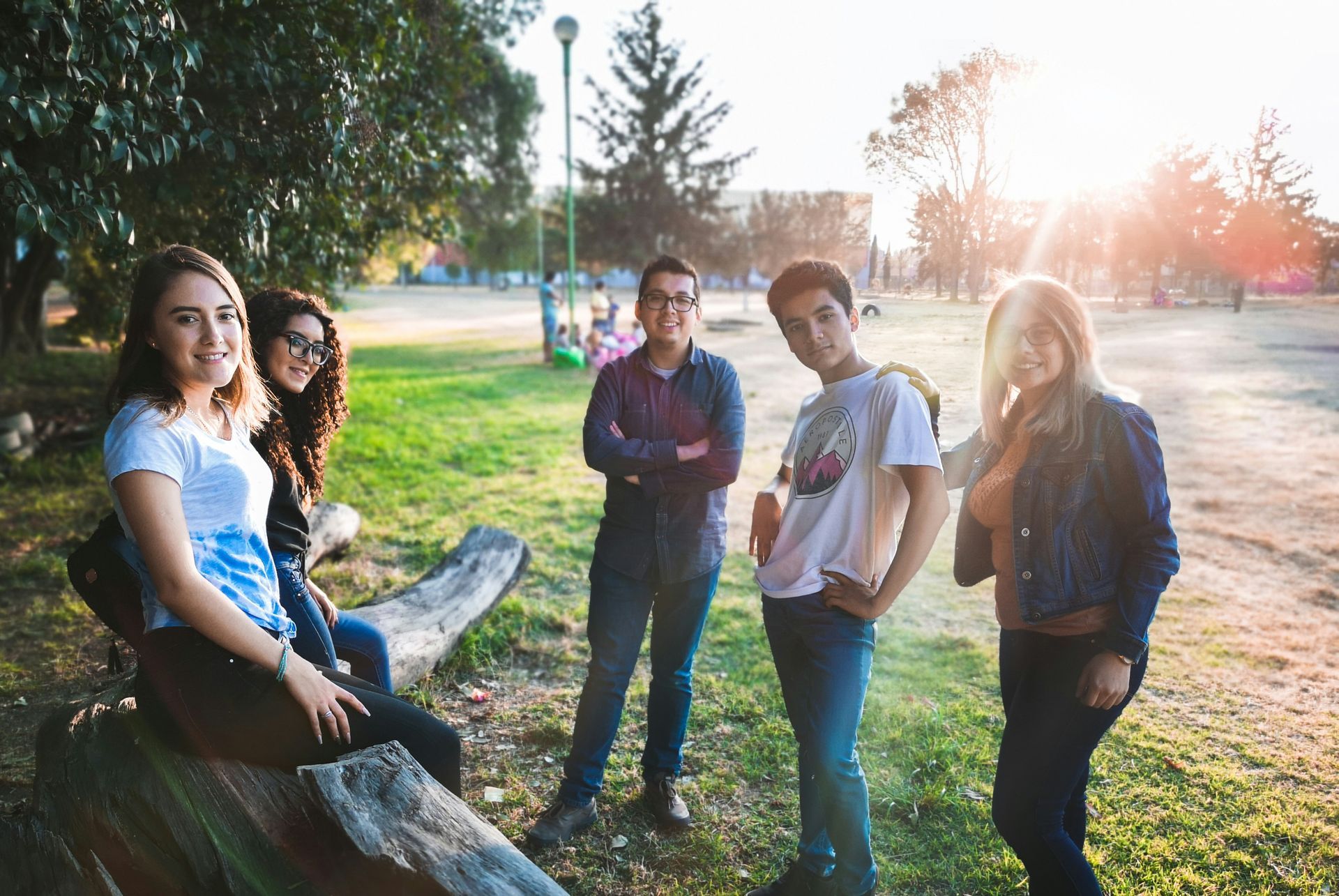 Five people pose outdoors on a sunny day in a grassy park, some sitting on a log and others standing.