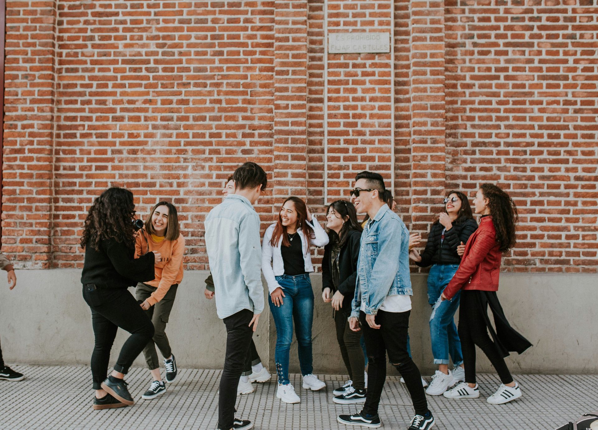 A group of young people laughing and chatting while standing together in front of a red brick wall.
