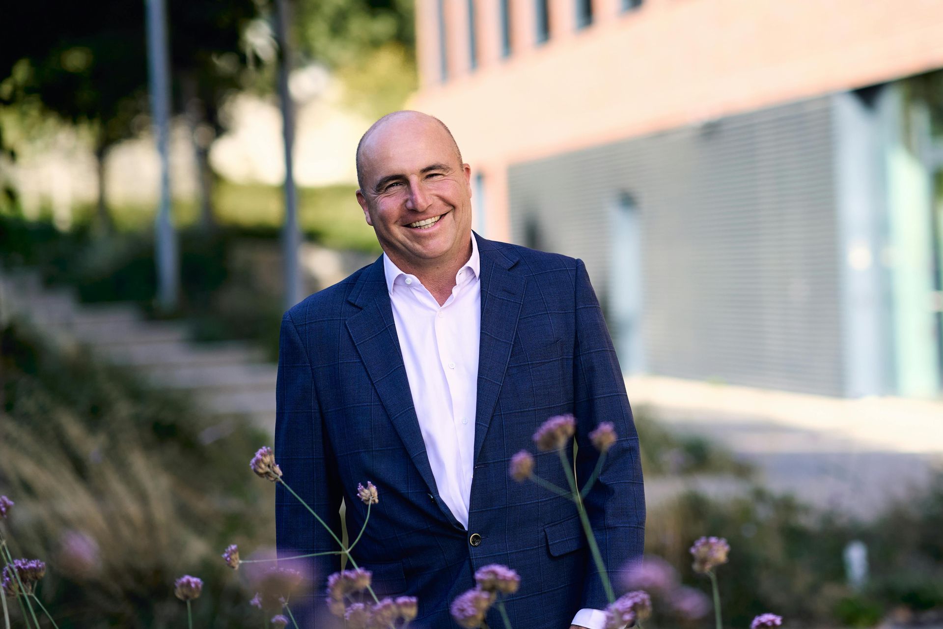 A smiling bald man in a white shirt and navy blazer standing outdoors in front of a building with purple wildflowers.