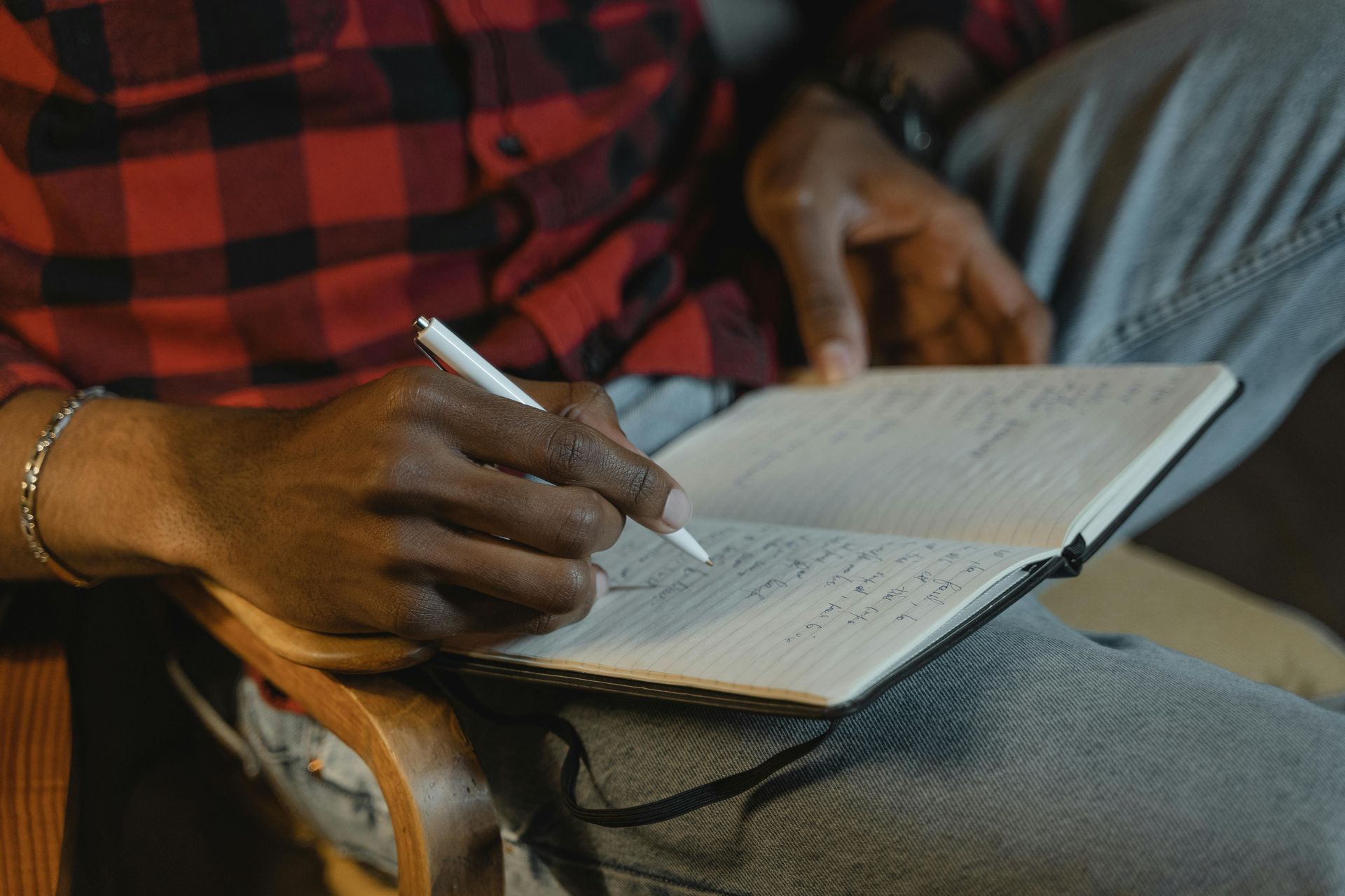 A person wearing a red plaid shirt writes with a white pen in a lined notebook while sitting in a wooden chair.