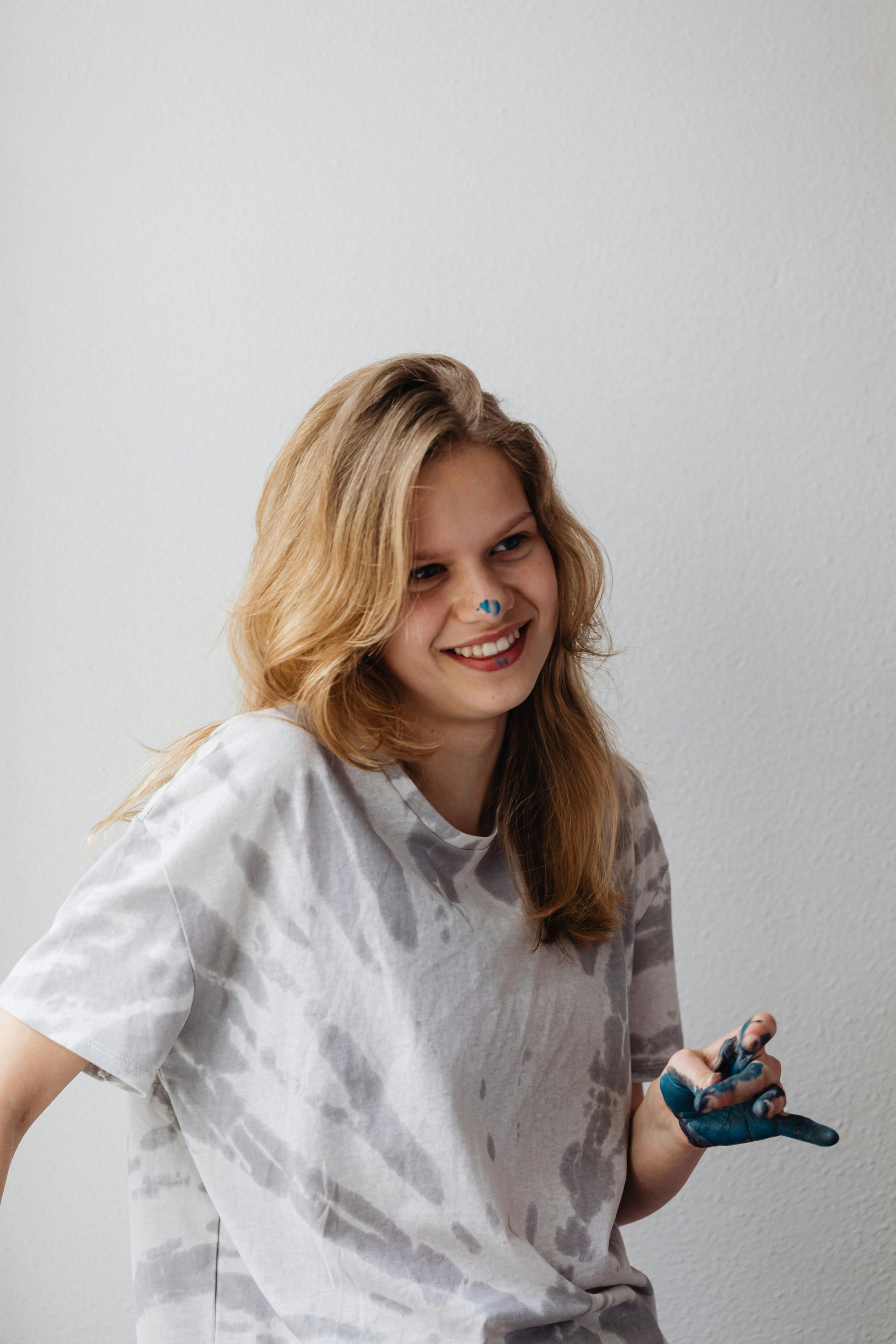 A person with blonde hair and a blue-stained hand smiles, wearing a tie-dye shirt against a plain white background.