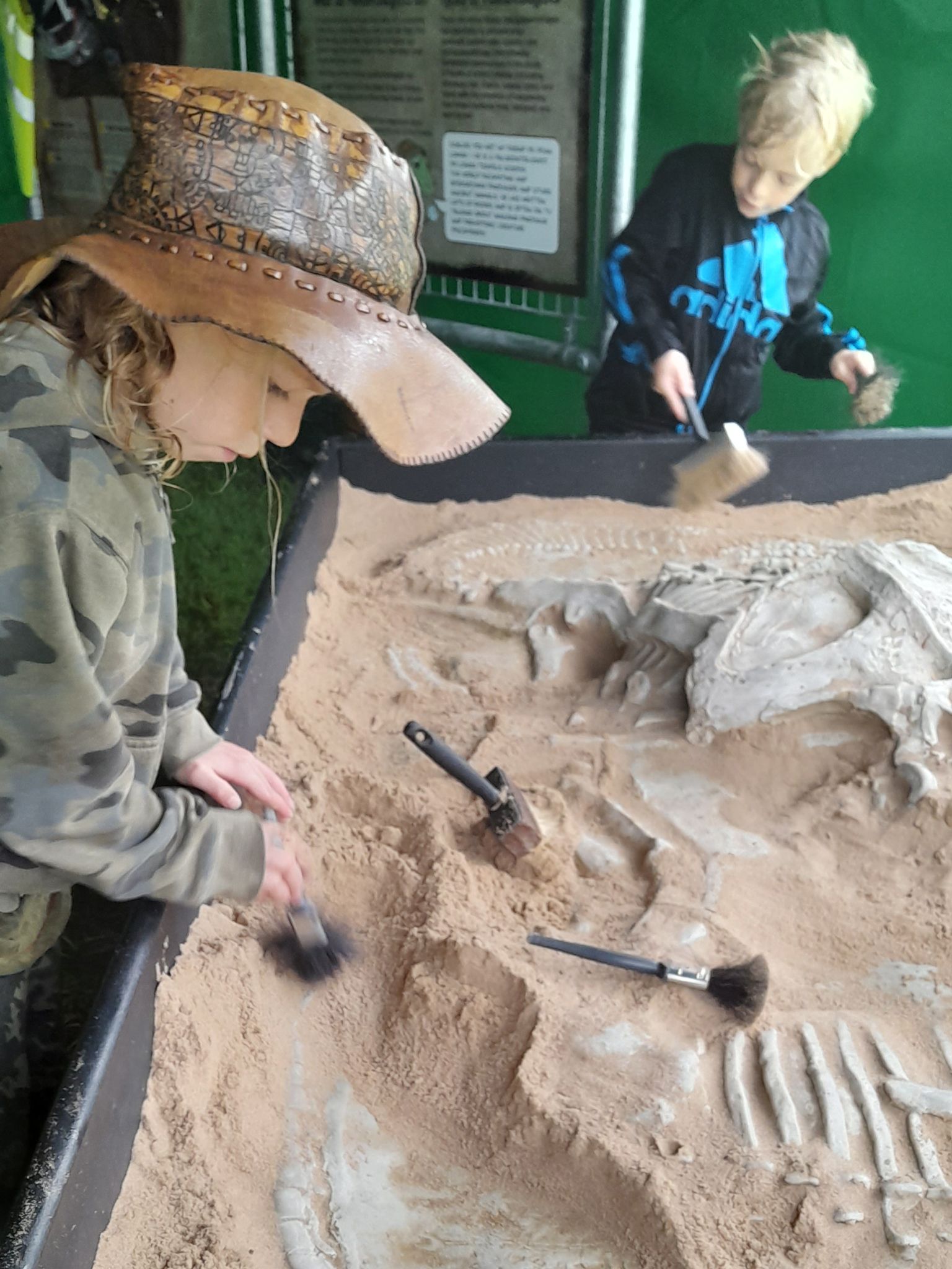 Two young boys are digging in the sand near a dinosaur skeleton.