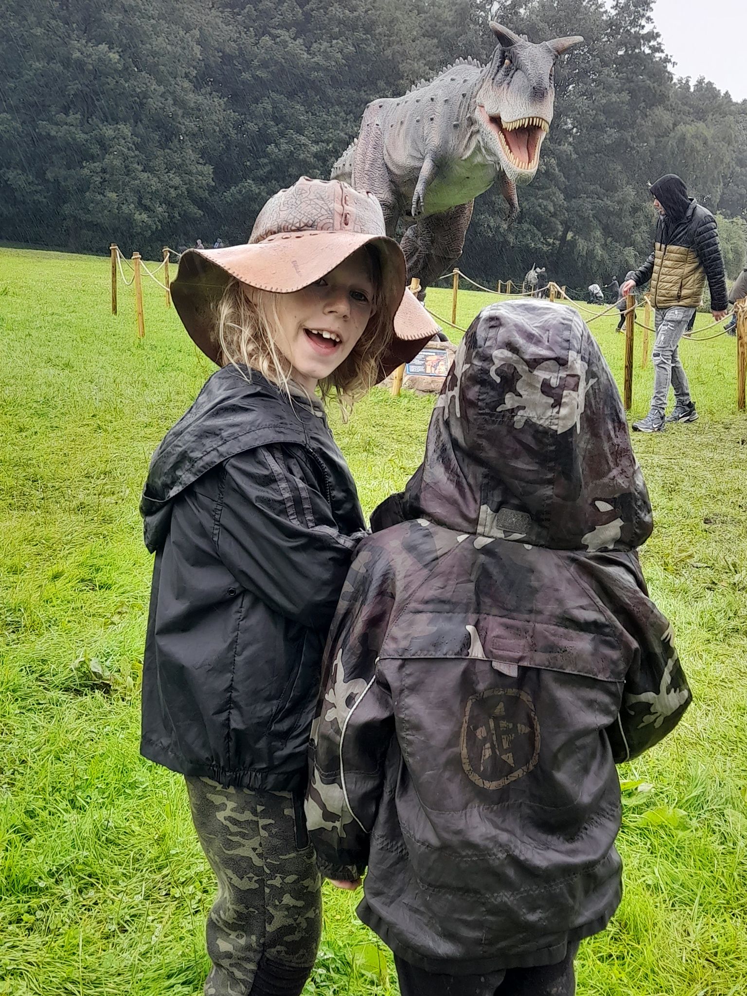 Two children are standing next to a dinosaur statue in a field.