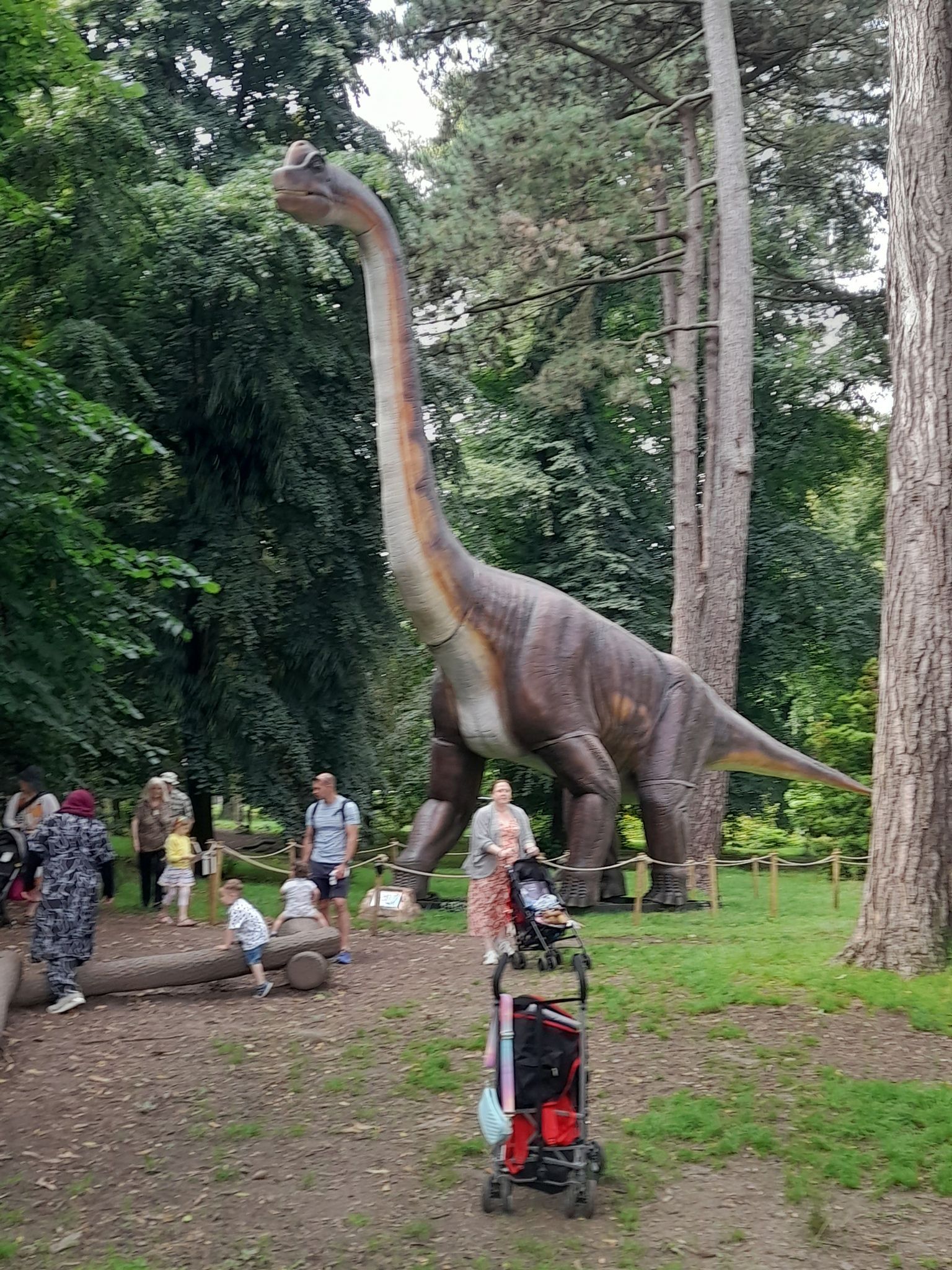 A statue of a dinosaur in a park with people standing around it.