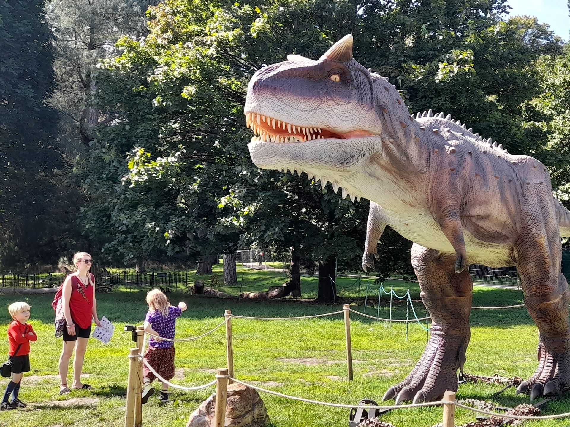 A large dinosaur model in a park with people standing around it.