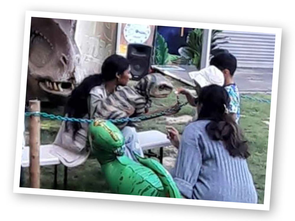 A group of people are sitting under a tent in front of a dinosaur.