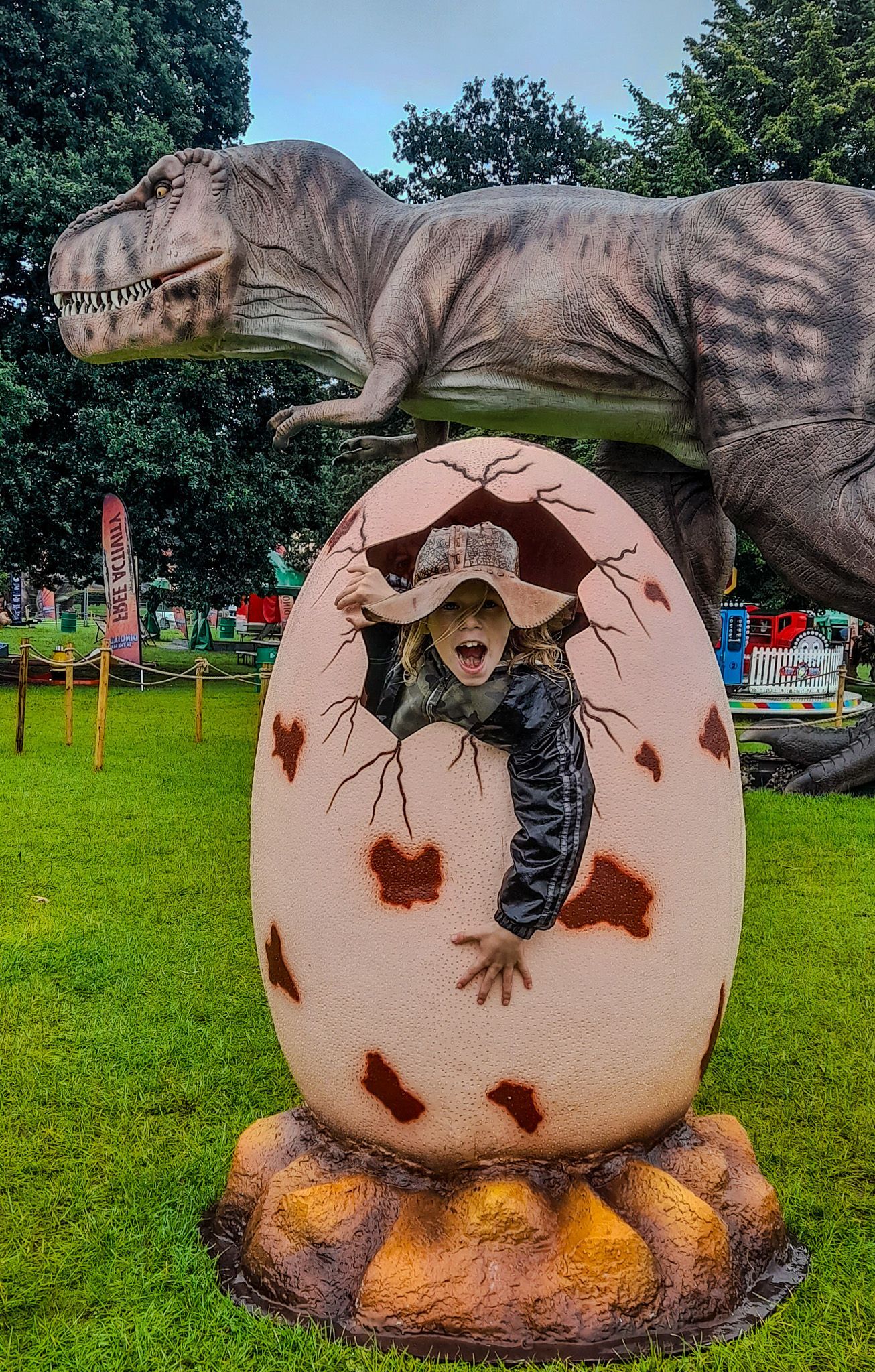 A child is coming out of an egg with a dinosaur statue in the background.