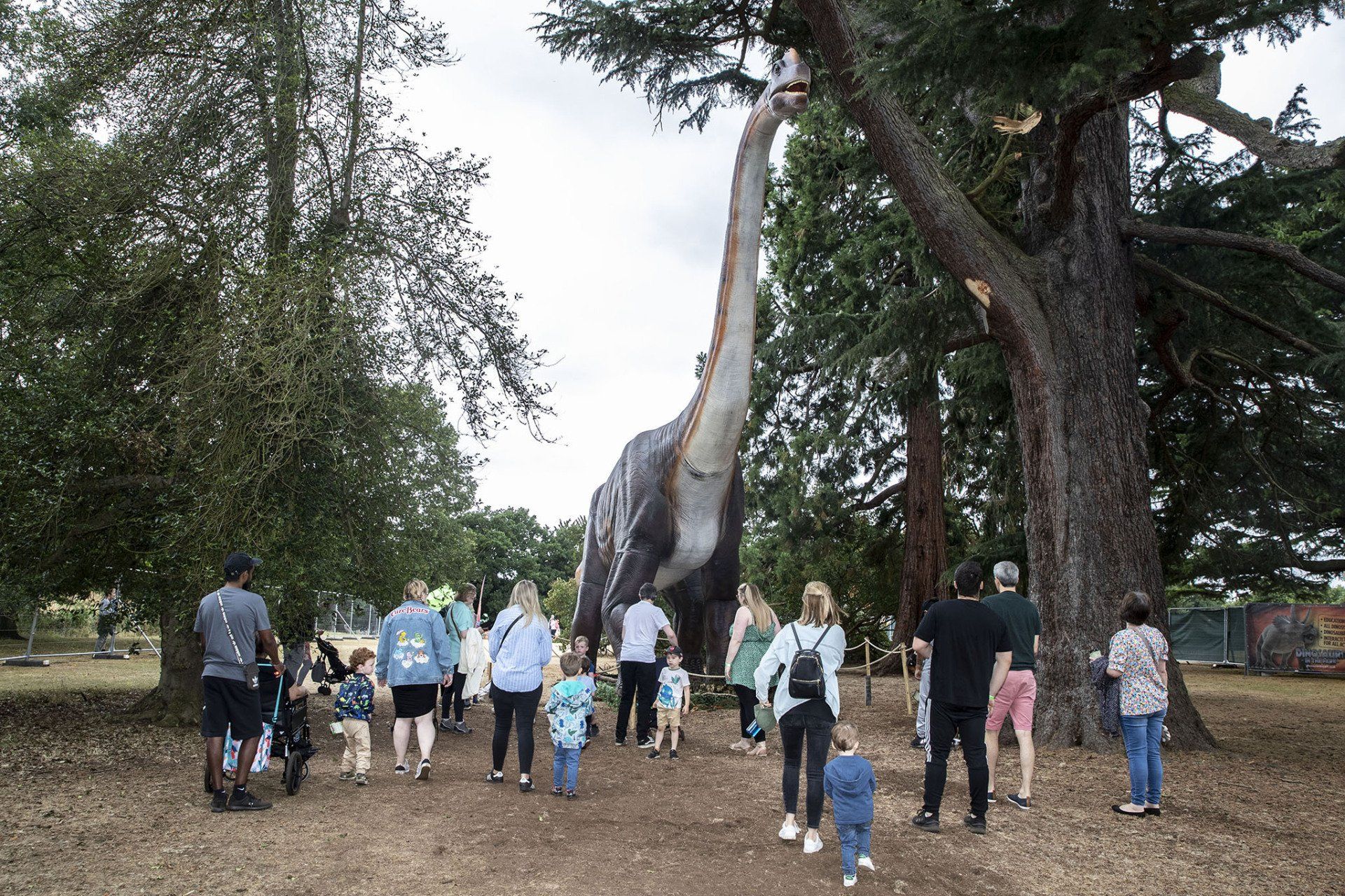 A group of people are walking in front of a large dinosaur model in a park.