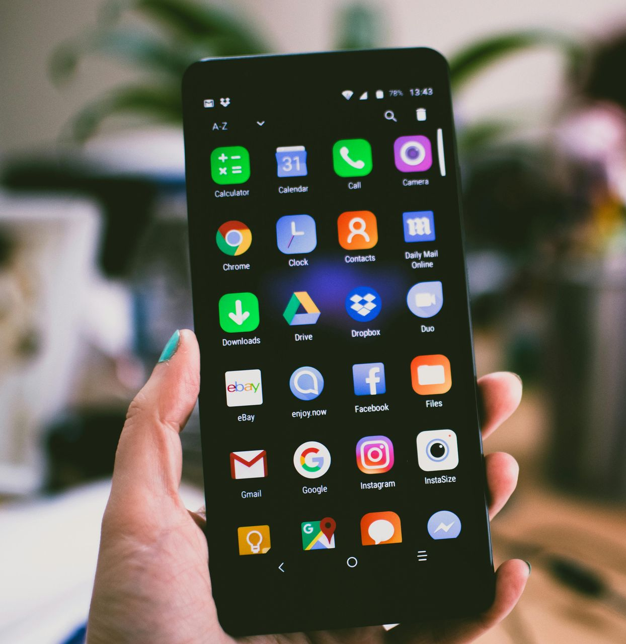 Hand holding black smartphone displaying app icons; desk with a laptop and plant in the background.