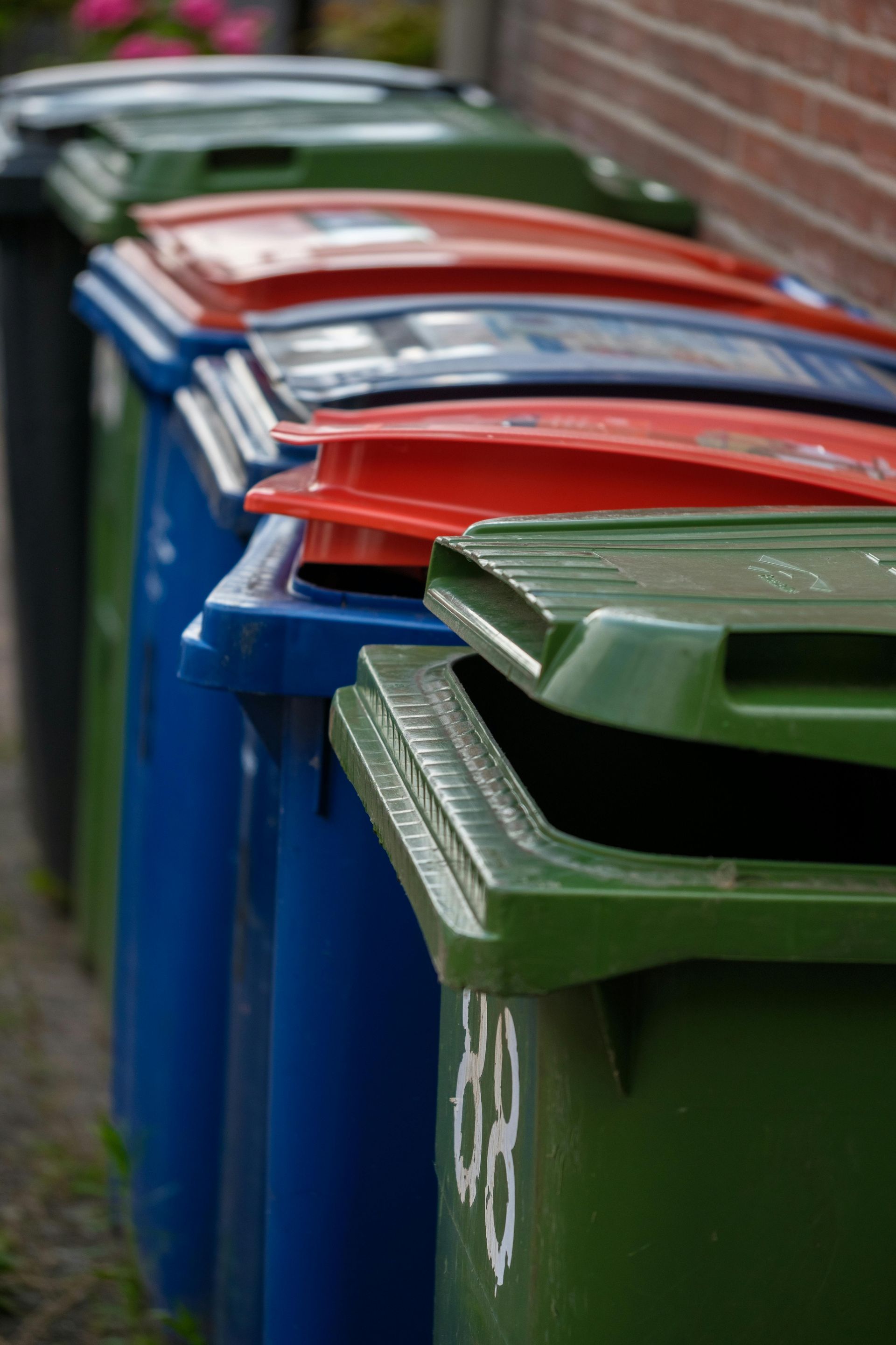 Row of colorful trash bins: green, blue, red. One green bin lid open, in front of a brick wall.