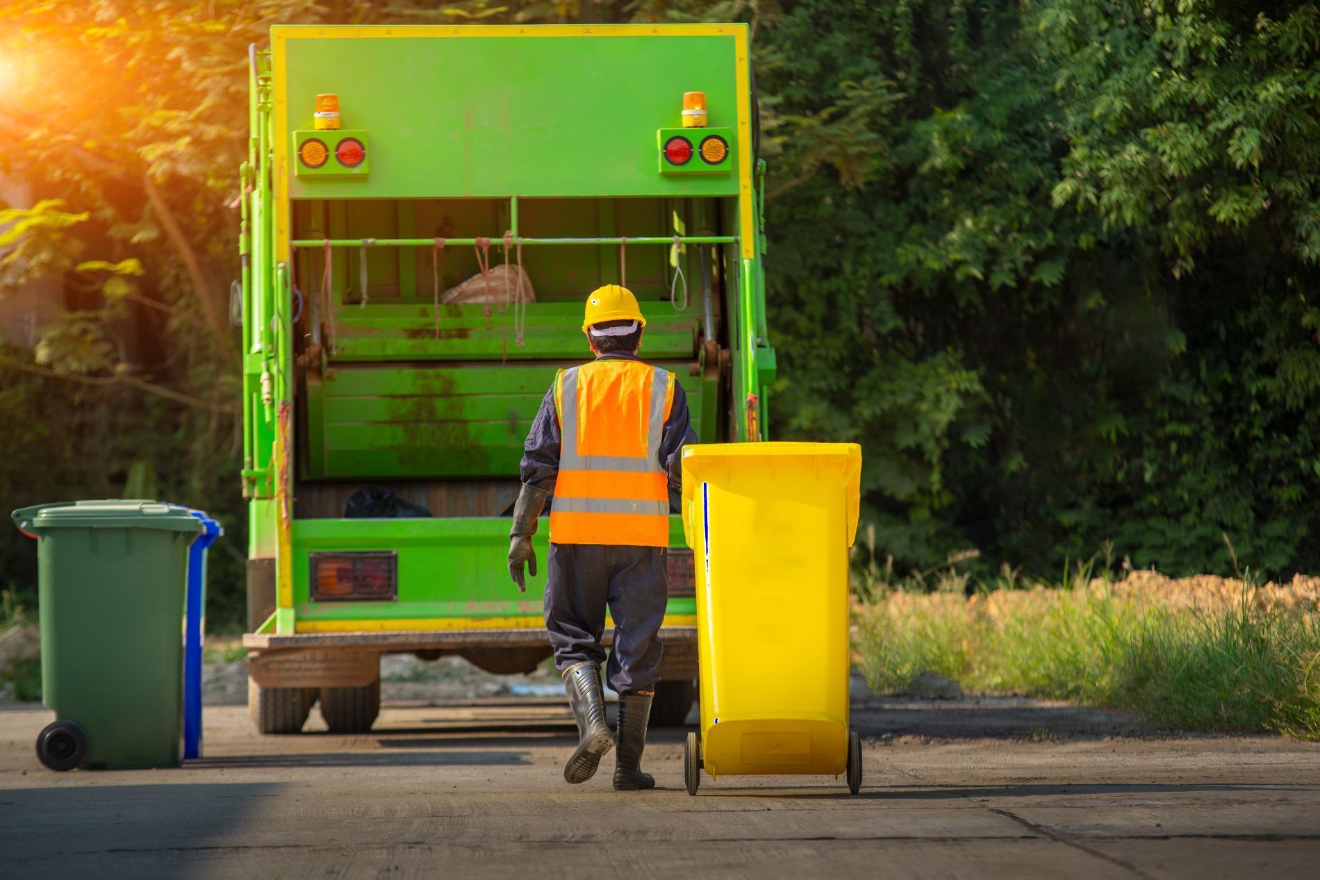 Garbage truck worker with safety vest empties a yellow trash bin into a green truck on a sunny road.