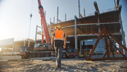 Construction worker in orange vest walking toward a crane at a building site at sunrise
