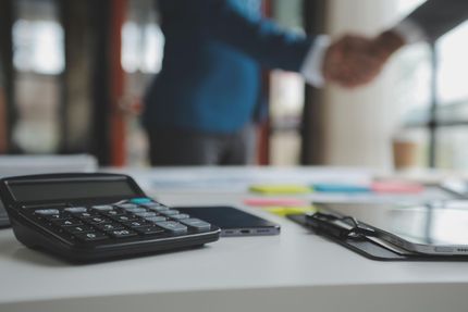 Calculator, smartphone, and glasses on a desk with two people shaking hands in the blurred background
