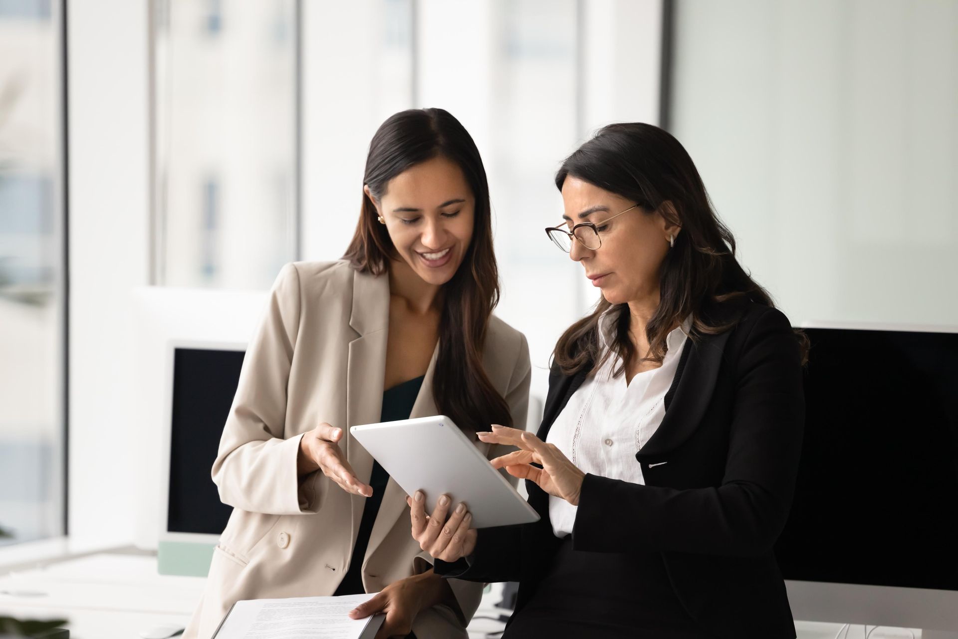 Two coworkers reviewing a document together in a bright office, smiling and pointing at a tablet.