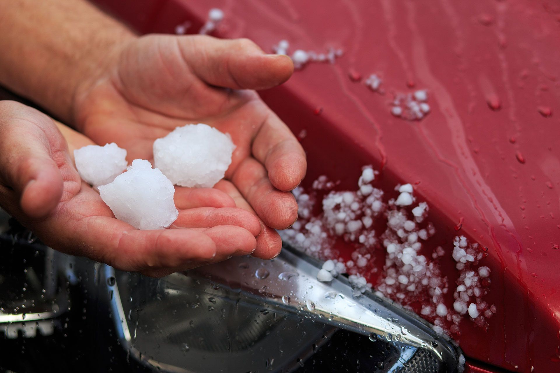 Hands holding hailstones on a red car hood, after a hailstorm.