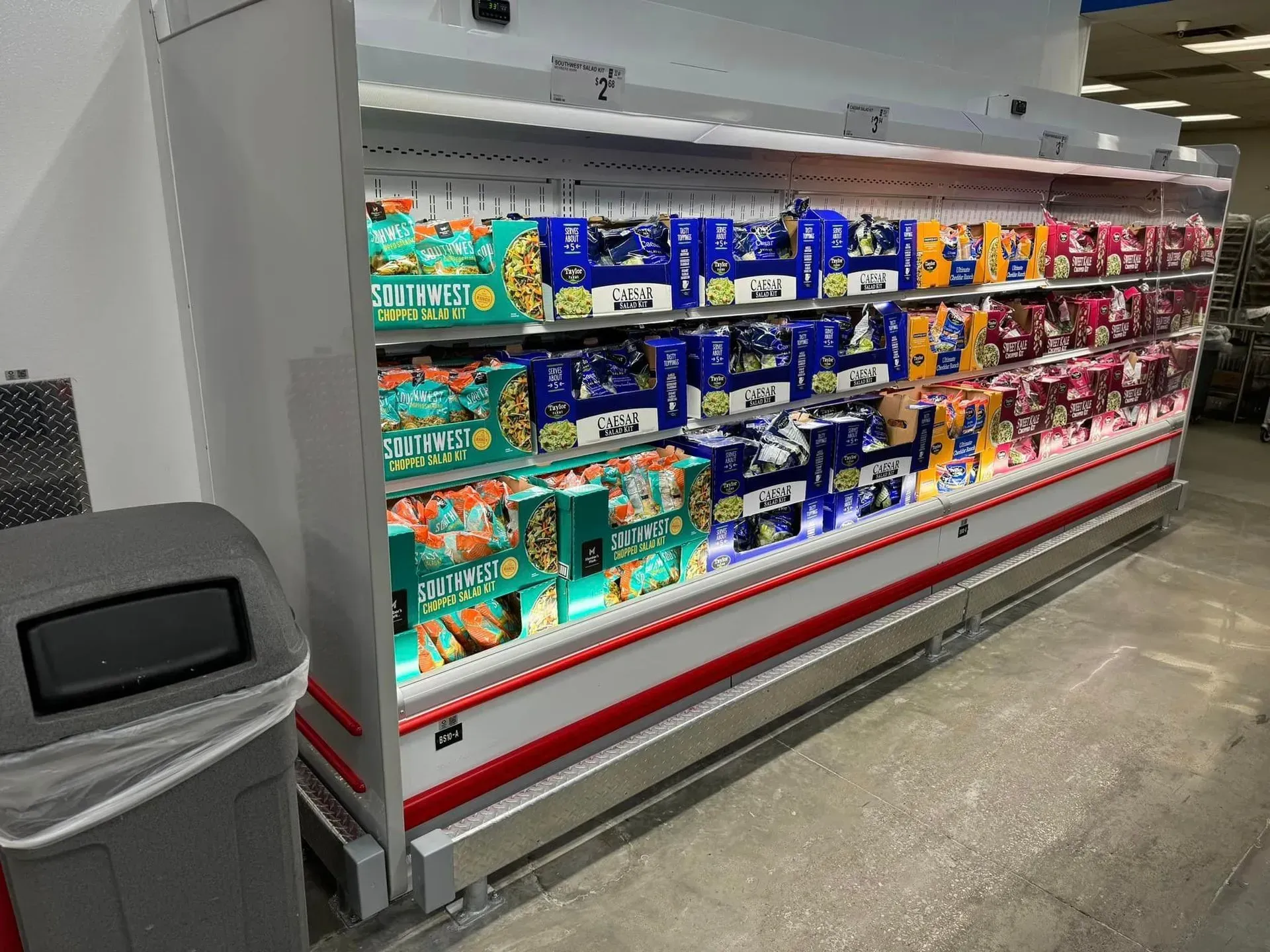 A refrigerator filled with lots of food in a grocery store next to a trash can.