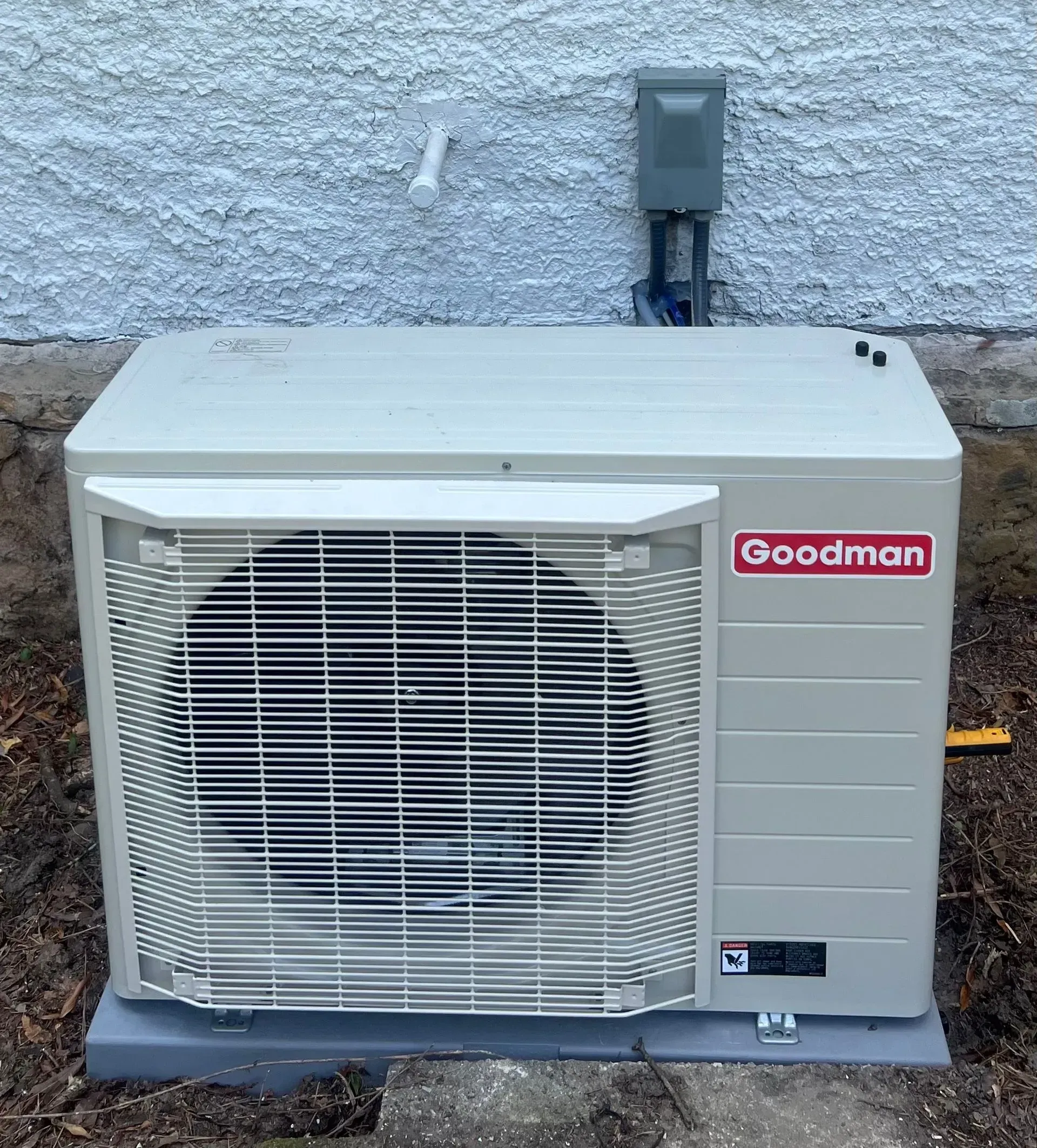 A goodman air conditioner is sitting on the ground next to a wall.