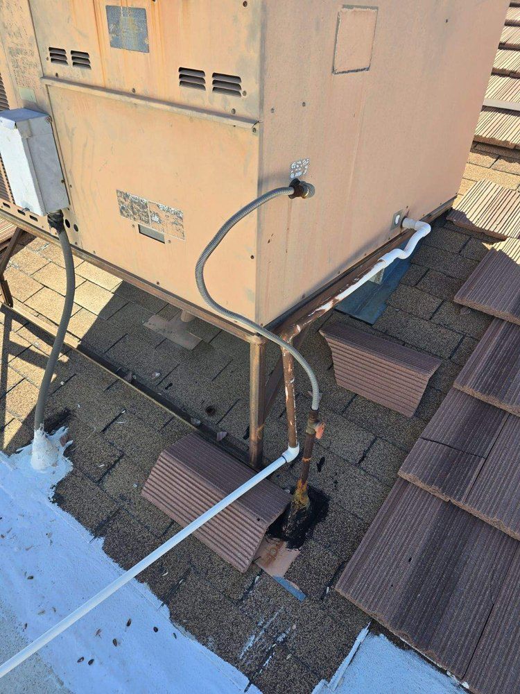 A rooftop HVAC unit with connected electrical conduit and water supply piping on a shingled roof.