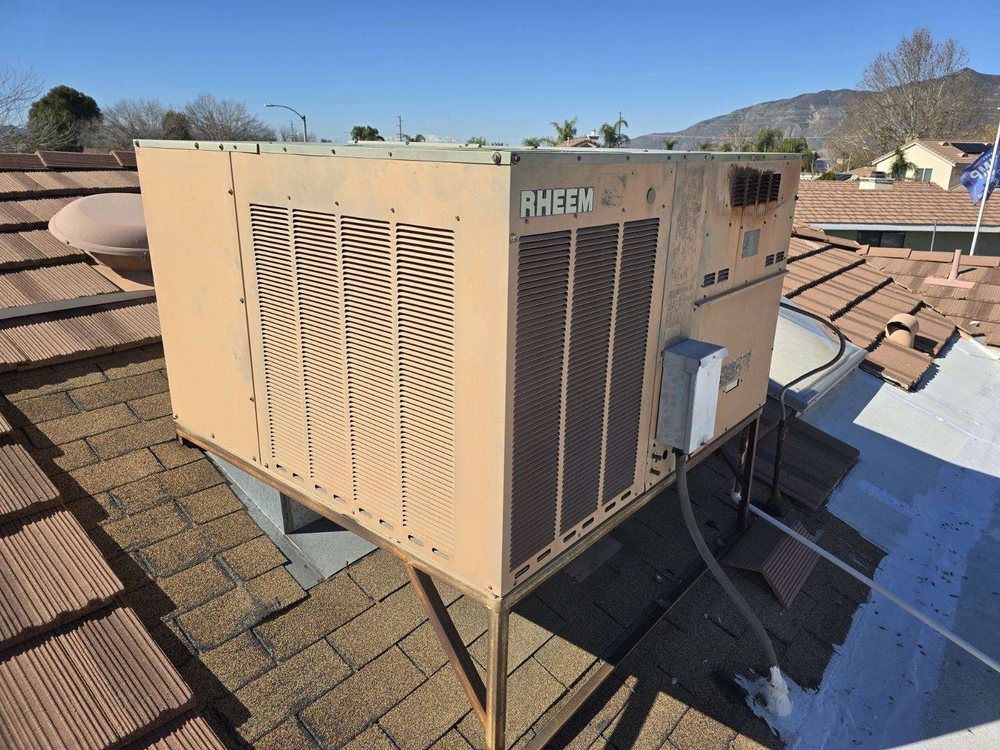A tan Rheem HVAC unit sits on a brown tiled roof under a clear blue sky, with a disconnect box wired to the side.