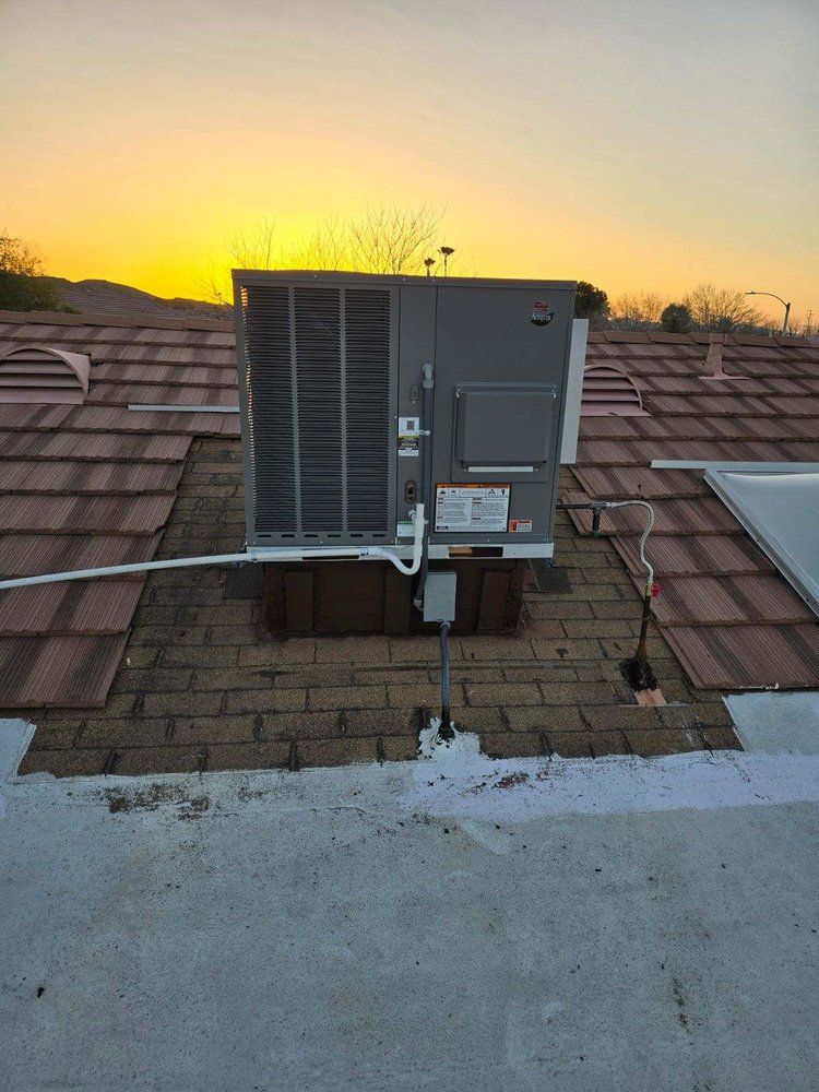 A gray HVAC unit sits on a tile roof at sunset, with a white conduit pipe leading to a disconnect box.