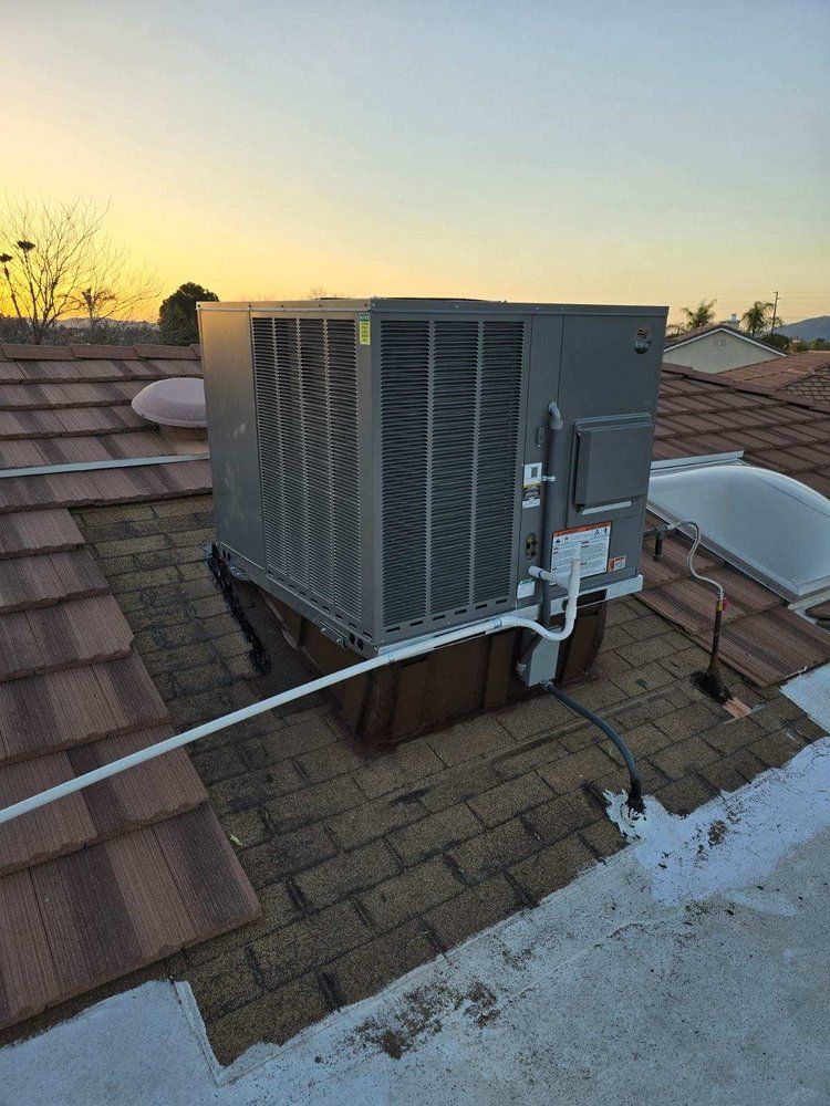 A gray HVAC unit sits on a residential rooftop with brown tile and shingle sections during sunset.