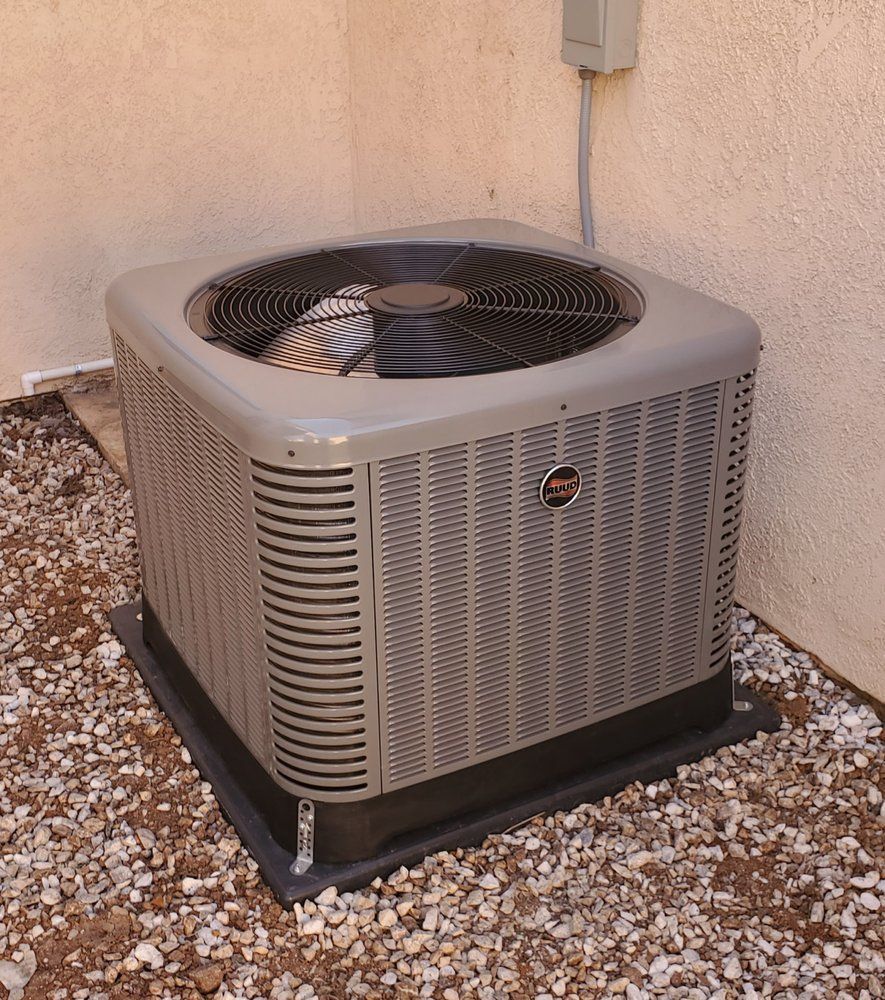 A grey residential HVAC condenser unit sits on a black pad atop gravel outside a beige stucco wall.