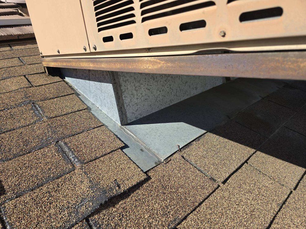 A tan HVAC unit sits on a roof, featuring a gray metal curb and flashing where it meets the brown asphalt shingles.