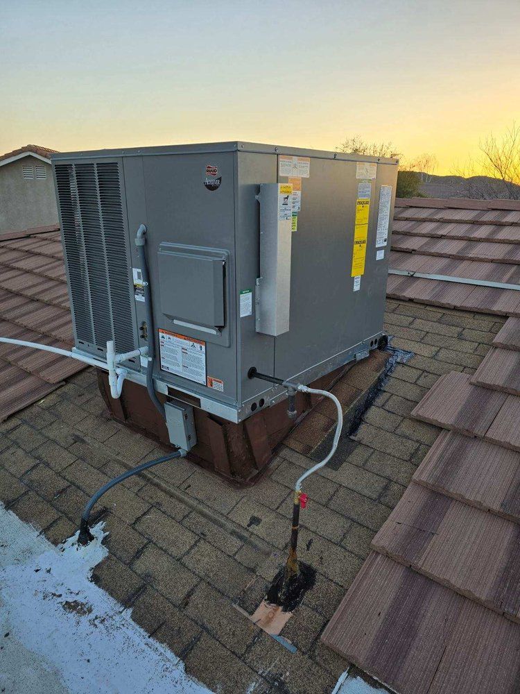 A grey HVAC rooftop package unit sits on a tile roof at sunset, featuring external electrical and gas line connections.