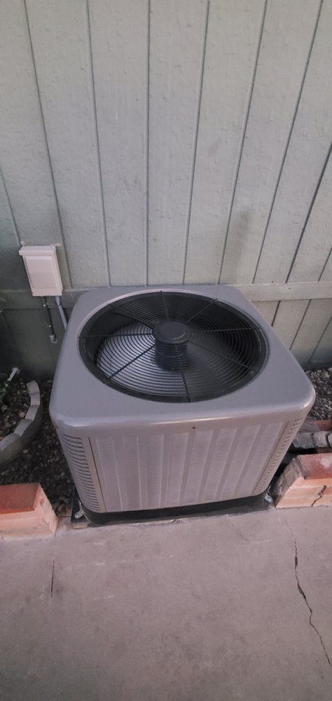 A square, grey outdoor HVAC condenser unit sits on a concrete pad against a light green wooden fence.