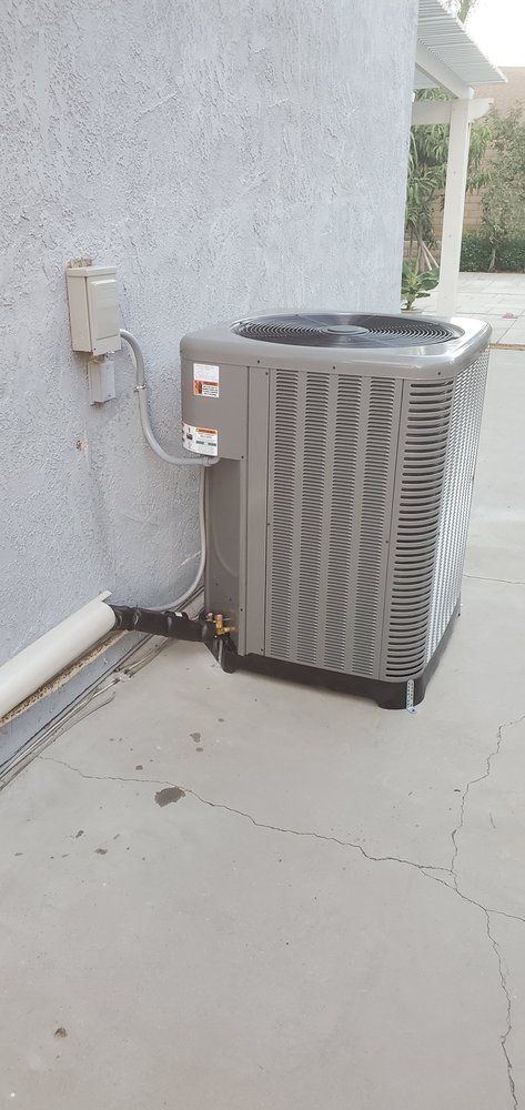A grey air conditioning unit sits on a concrete patio next to the light-colored stucco exterior wall of a house.