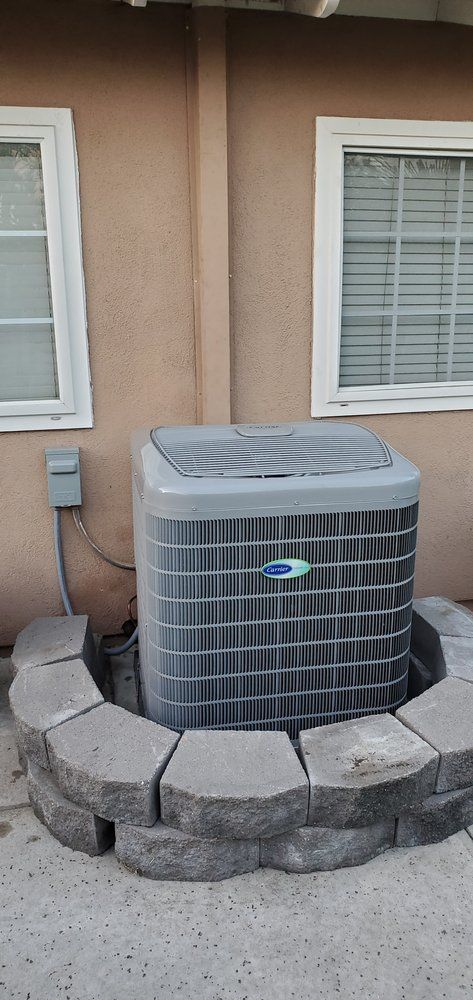 A gray HVAC unit sits on a patio against a tan wall between two windows, partially surrounded by a curved stone border.