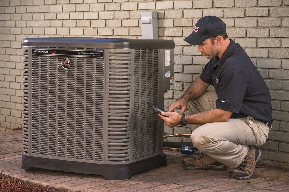 A technician wearing a work uniform kneels to inspect an outdoor residential air conditioning unit against a brick wall.