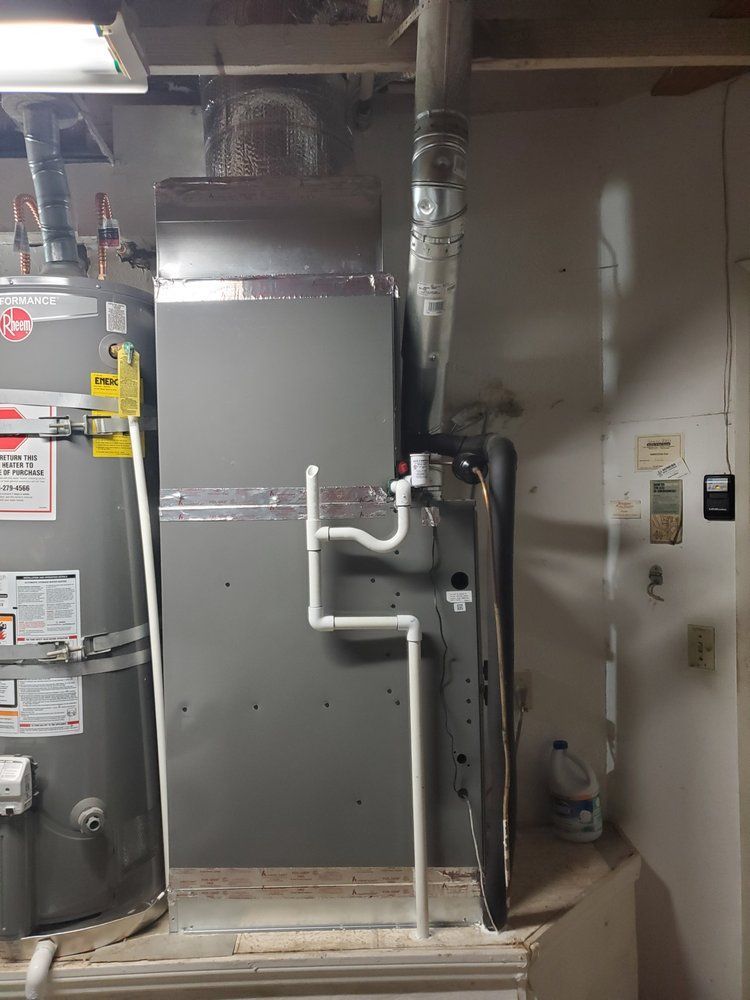 A gray HVAC furnace unit sits beside a water heater in a utility area, with white PVC condensate drainage pipes attached.