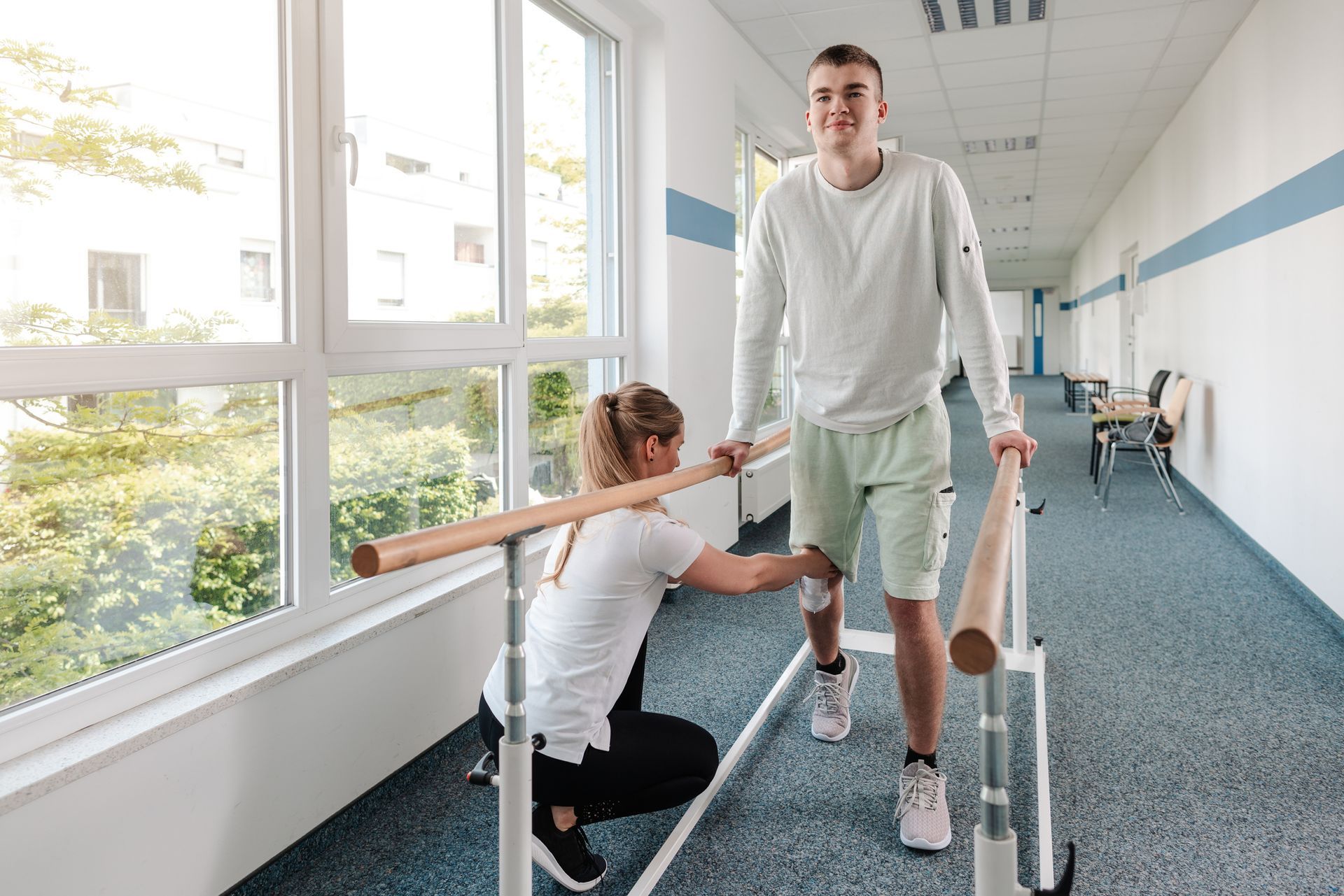A young man walks using parallel bars with assistance from a physical therapist in a brightly lit corridor.