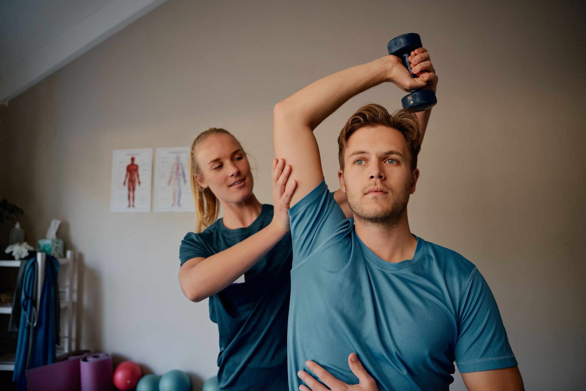 Physical therapist assists a man with arm exercise, holding a dumbbell overhead, in a rehabilitation setting.