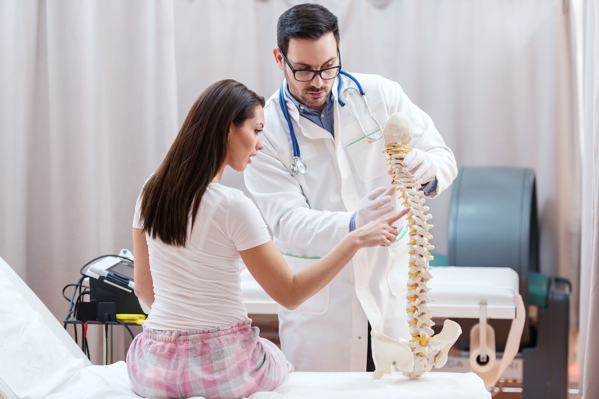 A patient sitting with her back turned and a doctor holding a spine model shows her the vertebrae.