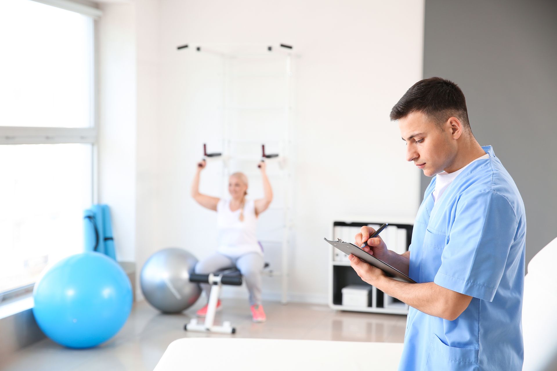 A physiotherapist writing notes in a rehabilitation centre with a patient on an exercise machine.