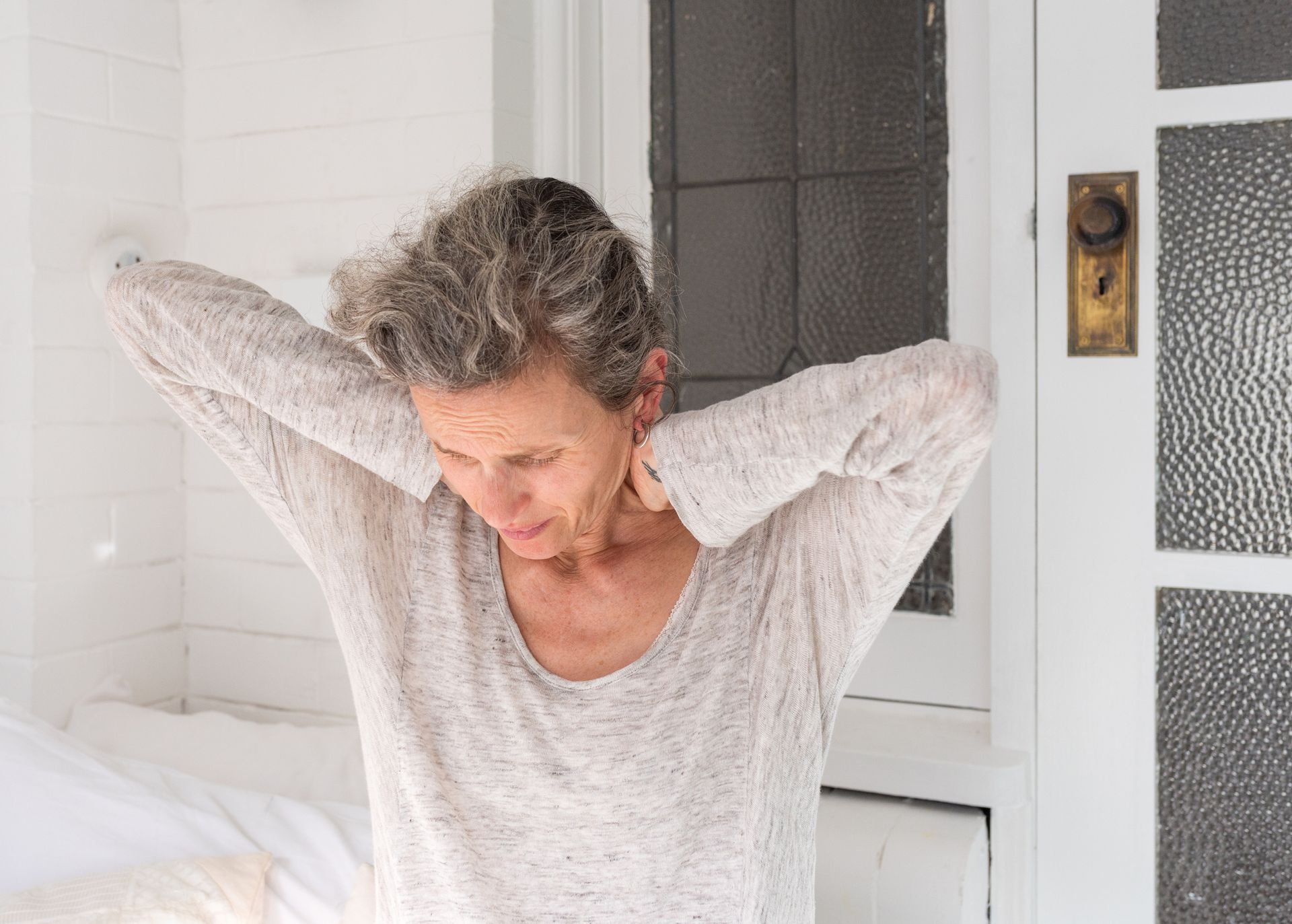 Woman with gray hair and a light sweater stretches her neck outdoors, looking downward.