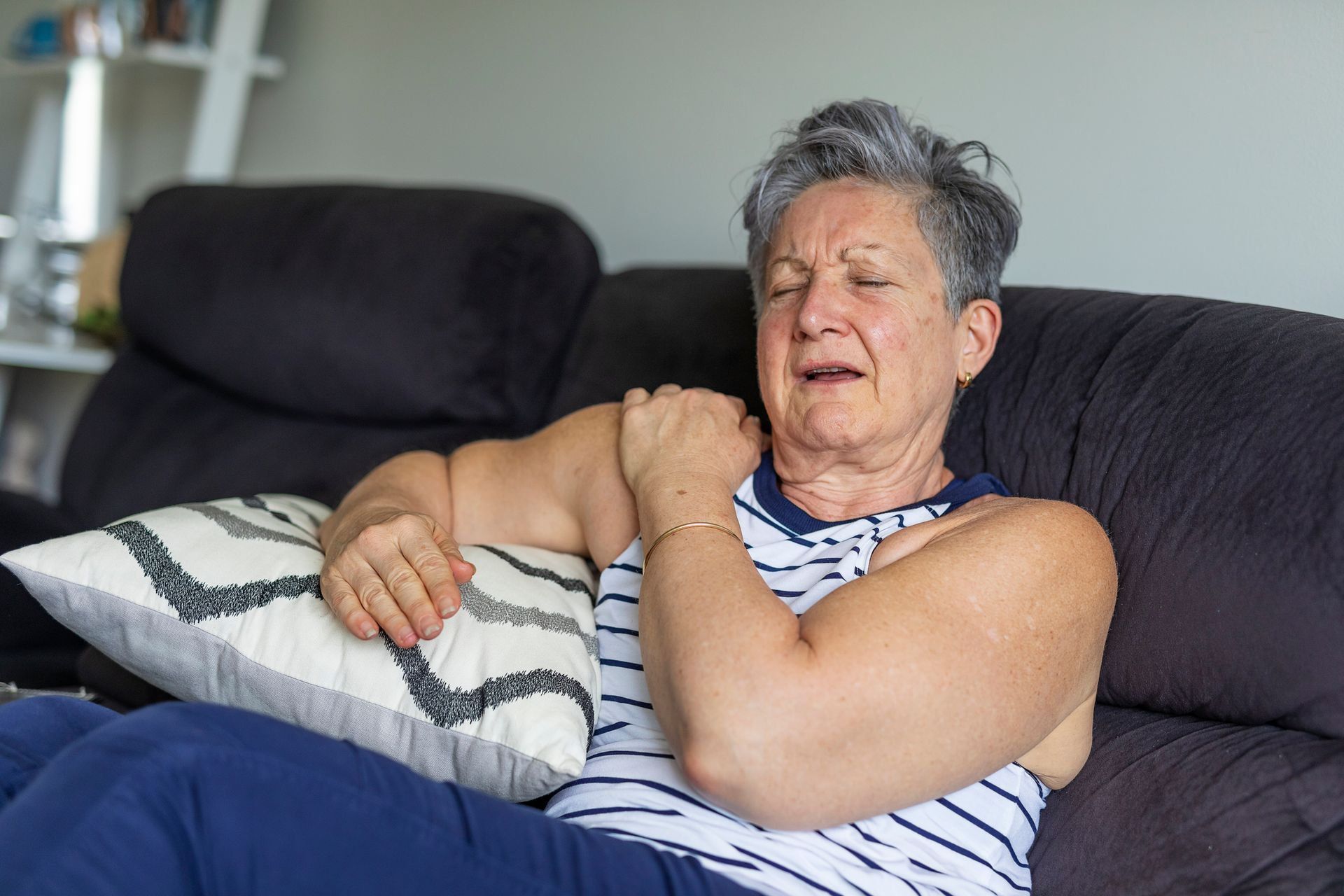 Older woman on a black couch, holding her shoulder in pain. She wears a striped tank top and looks distressed.