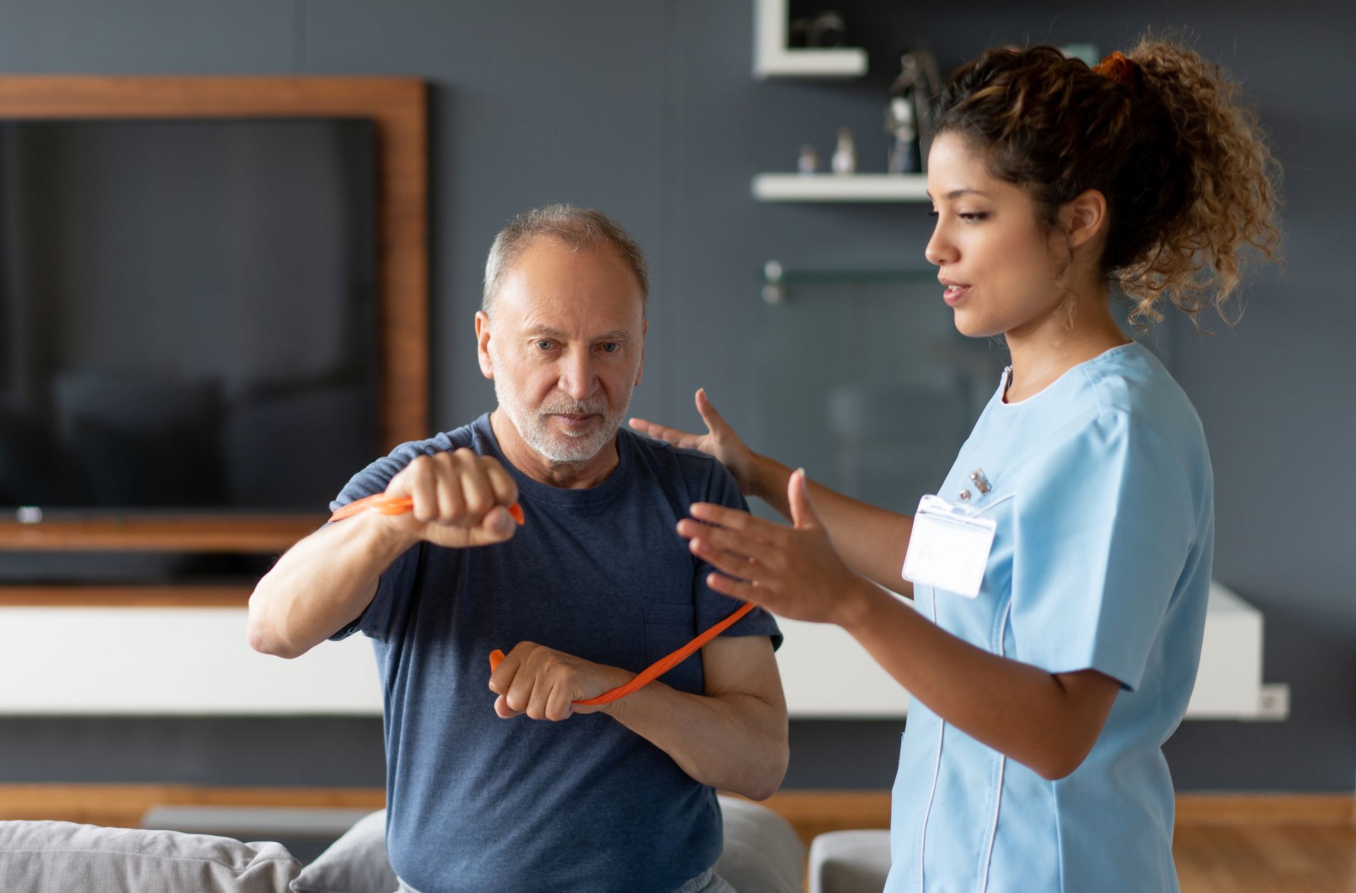 A physical therapist guides an older man using a resistance band for arm exercises in a living room.