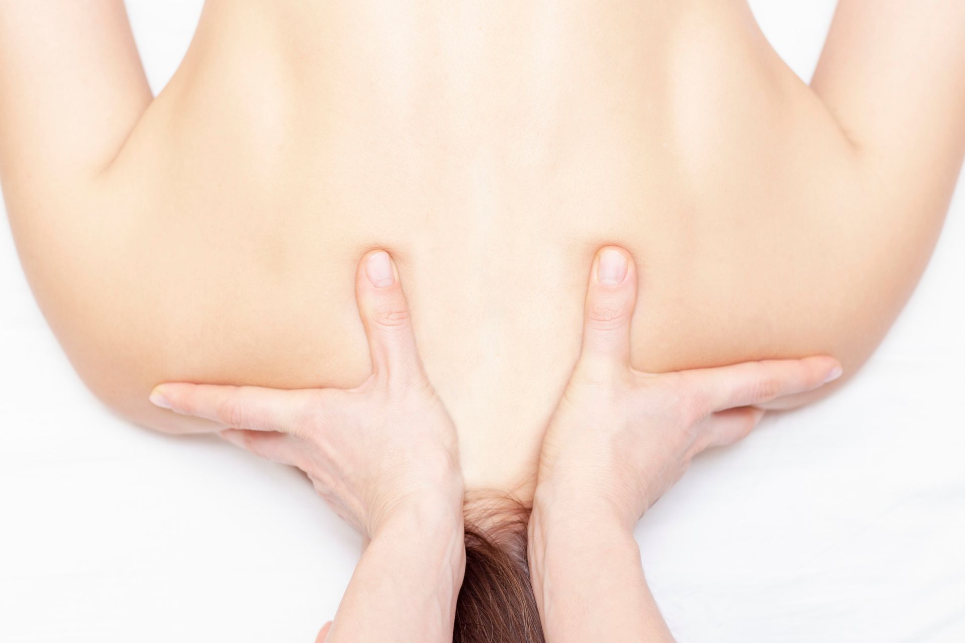 Hands massaging a person's upper back and shoulders. The person is face down on a white surface, skin-tone is pale.