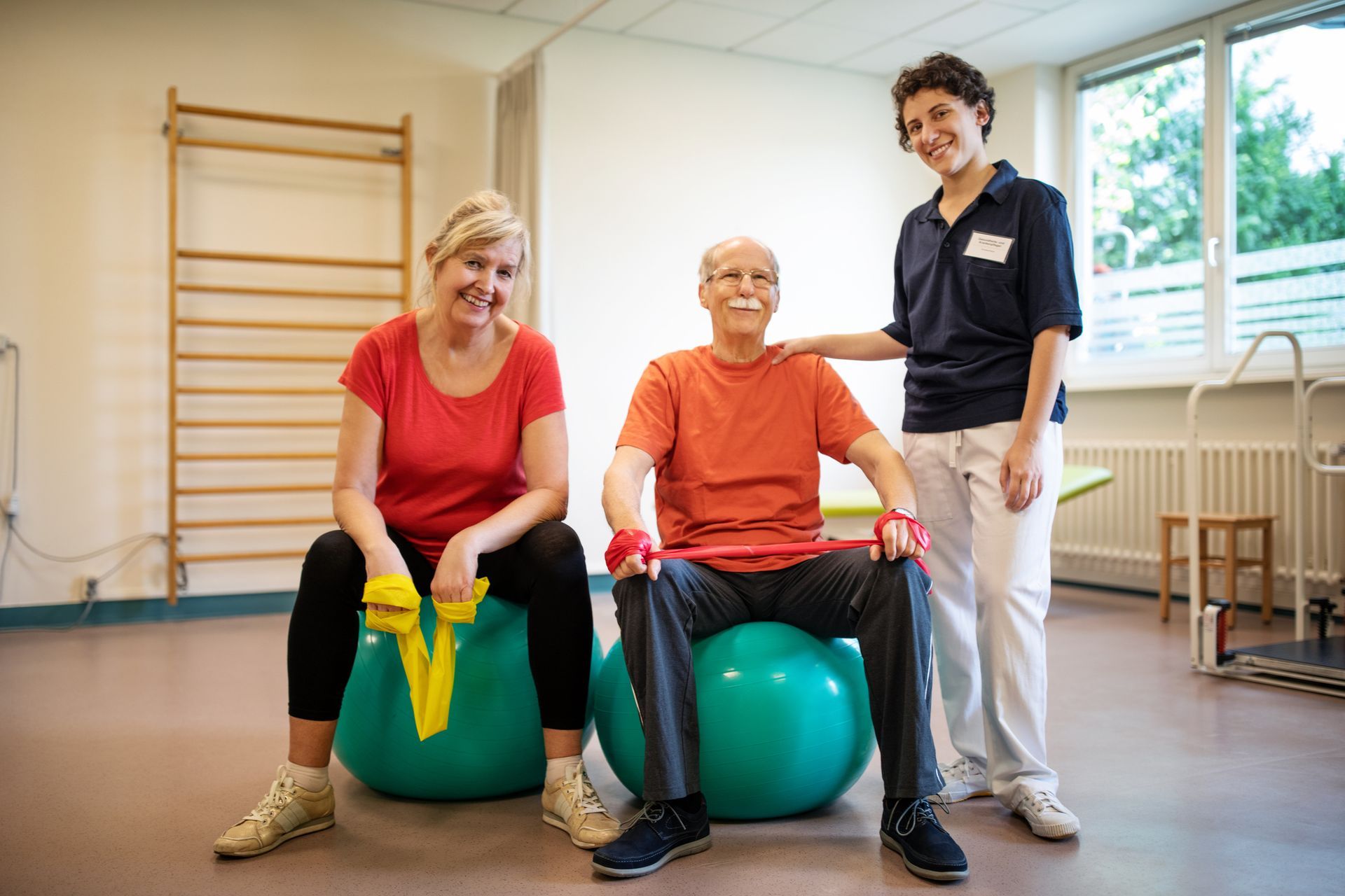 Two people and a therapist seated on exercise balls in a rehab room. The people smile, holding resistance bands.
